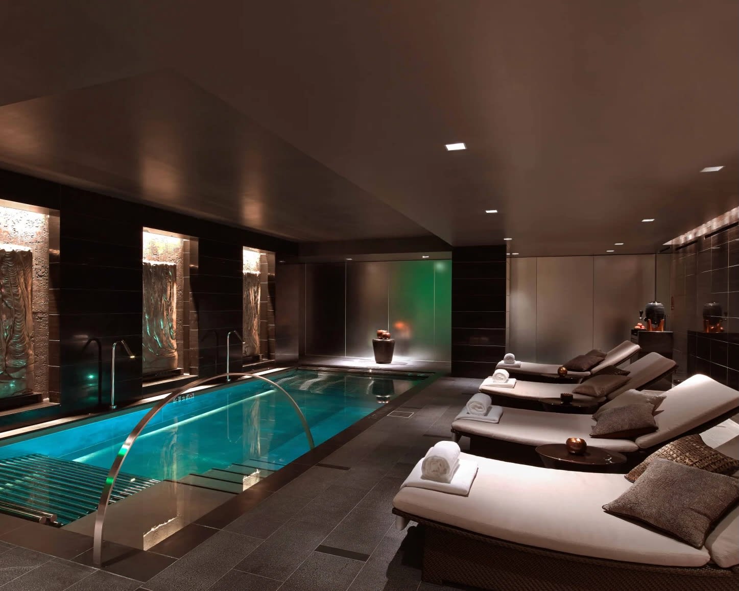 indoor dimly lit room with white lounge chairs next to a metal trimmed lap pool and dark walls
