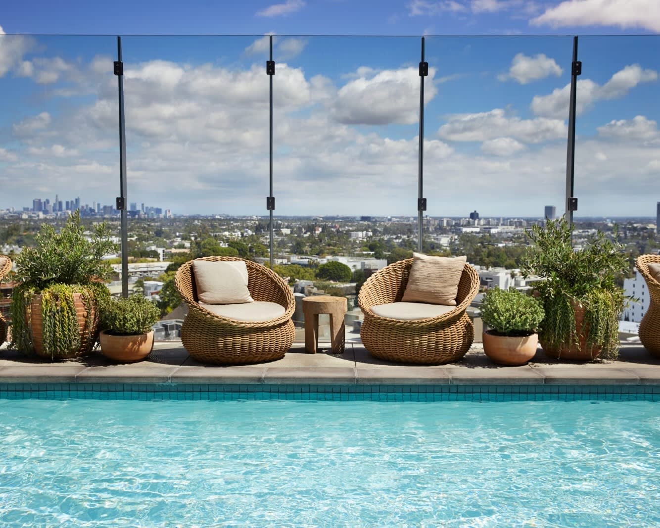 two circular wooden chairs next to a pool, surrounded by potted plants, and backed against a glass wall overlooking a city with skyscrapers in the distance during day
