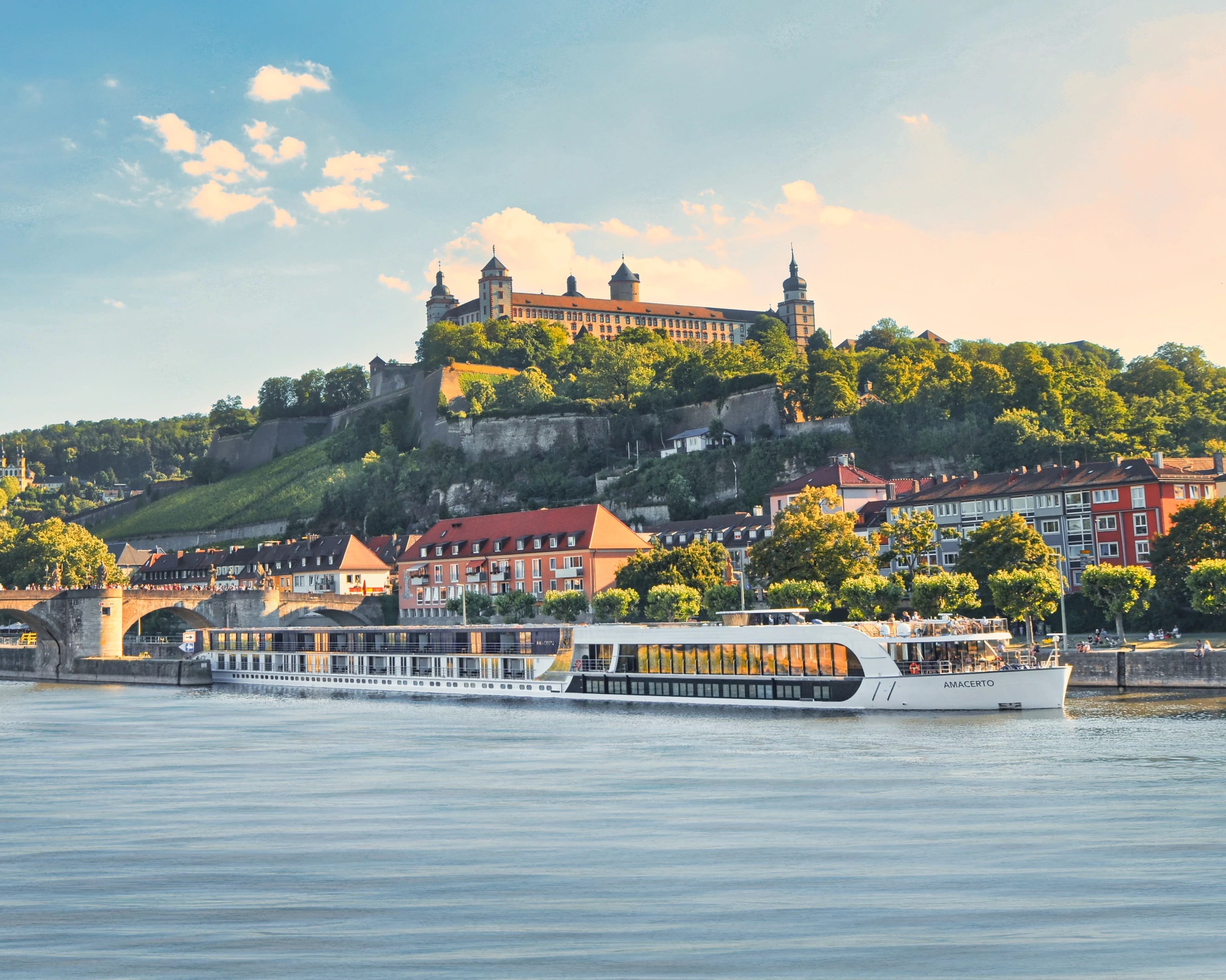 a white river cruise sailing along a river, under a bridge, with a hilltop castle behind it during day