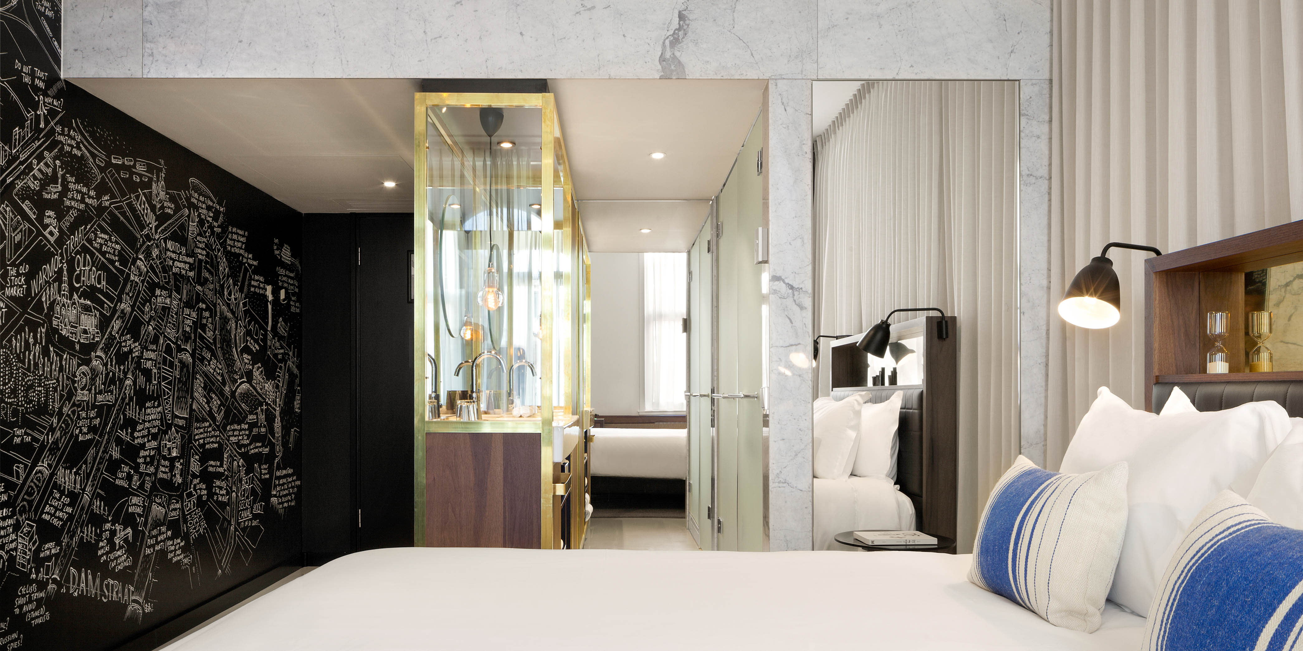 Contemporary hotel room with a black feature wall illustrated with a hand-drawn city map, glass-enclosed bathroom with brass detailing, and a neatly dressed bed with blue accent cushions.