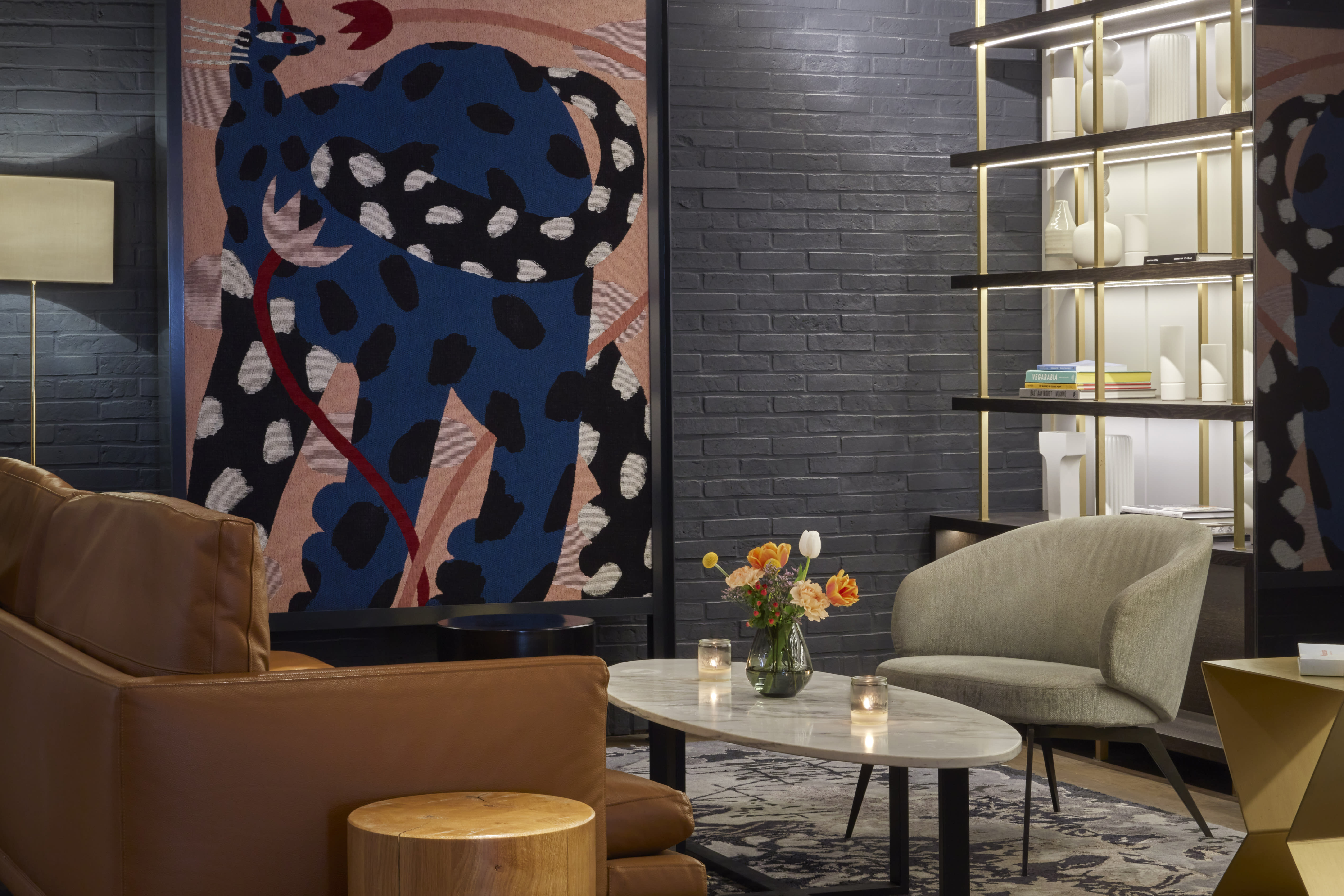 Contemporary lounge area with a bold abstract artwork in deep blue and pink tones, leather and upholstered armchairs, and a marble coffee table styled with candles and fresh flowers against a dark brick wall backdrop.