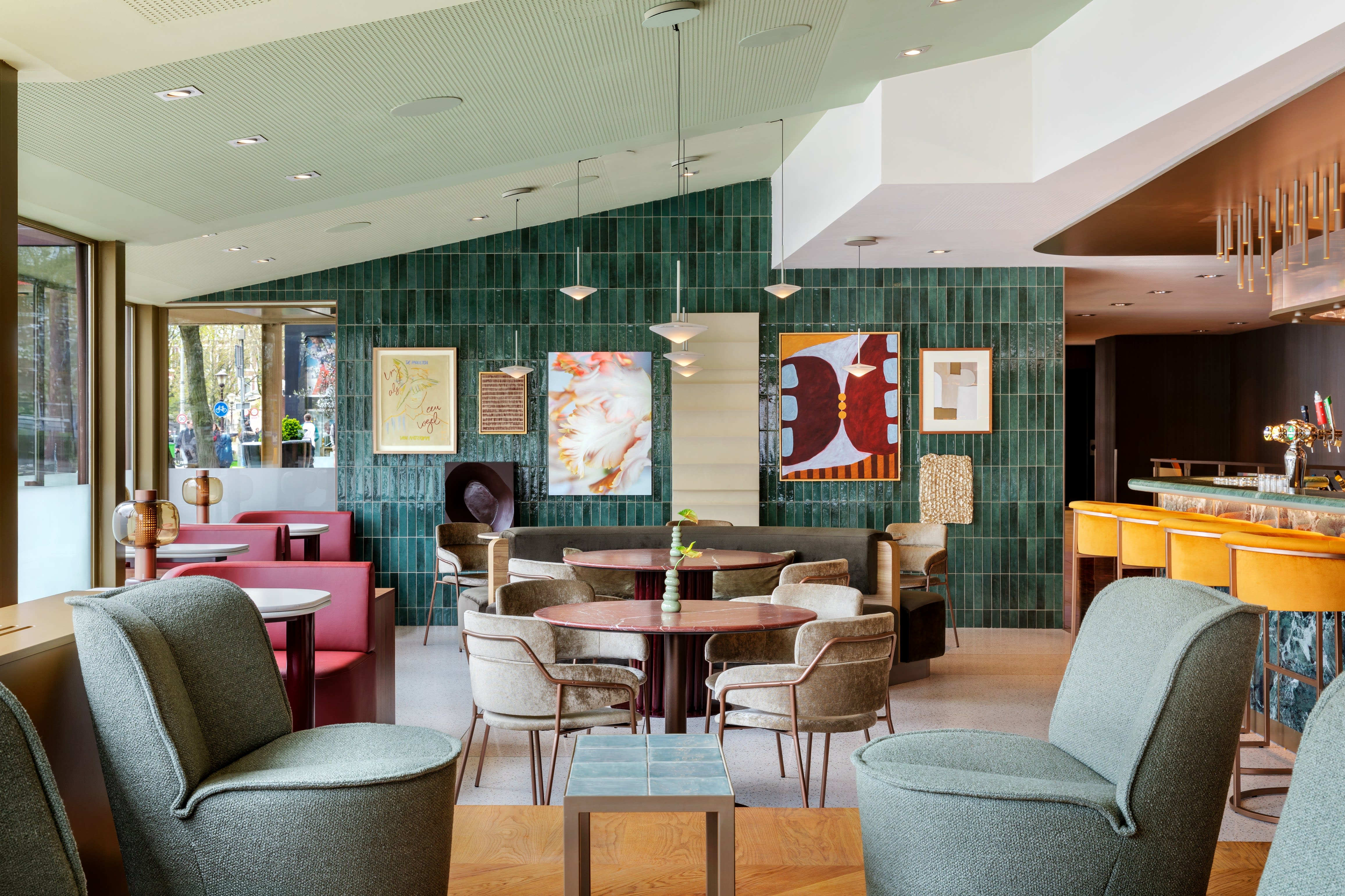 Stylish hotel bar and lounge with emerald green tiled walls, mid-century-inspired seating, and a curved bar lined with mustard velvet stools, accented by contemporary artwork and soft pendant lighting.