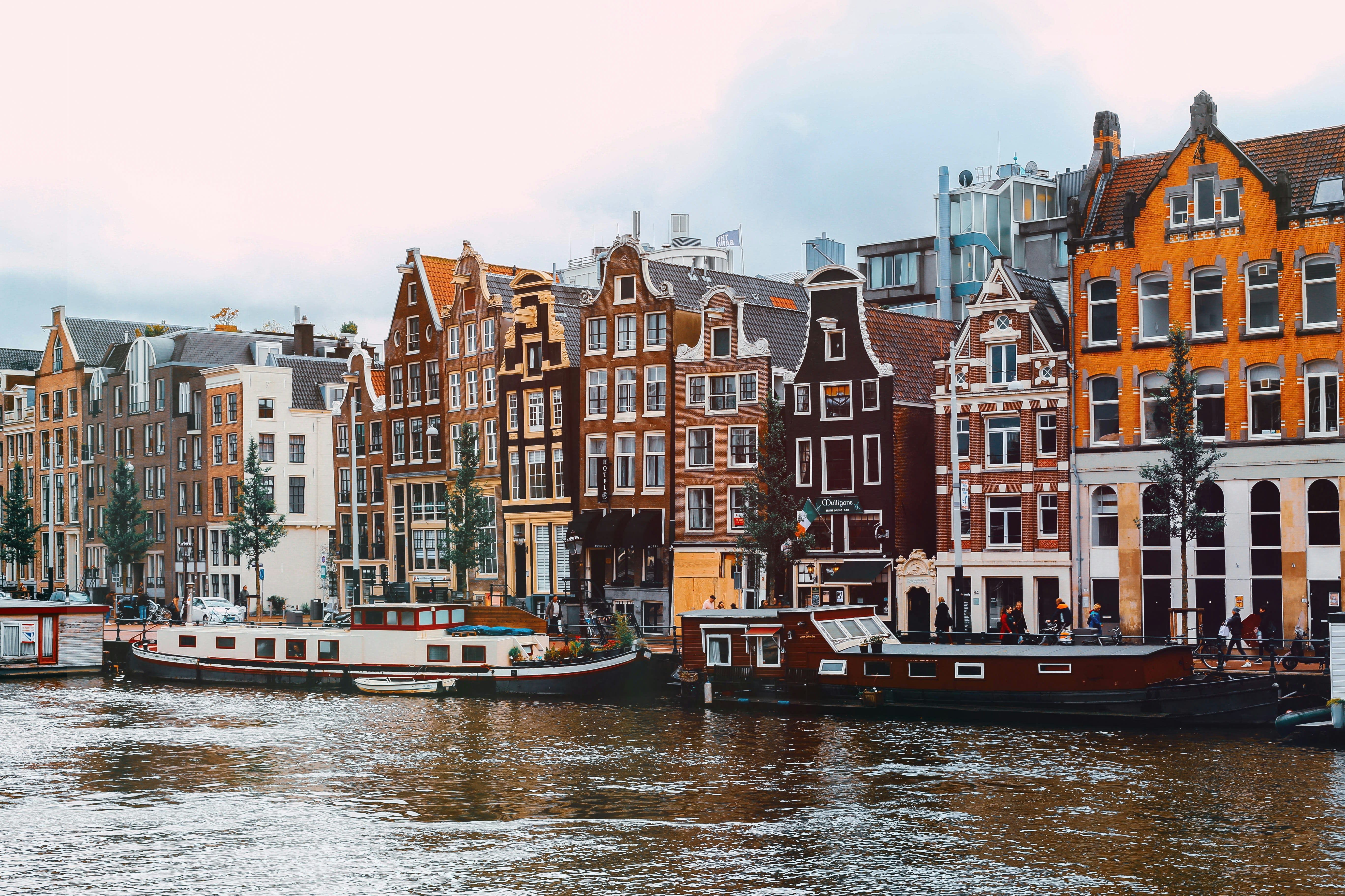 Row of traditional canal houses in Amsterdam with stepped gables and brick facades, lined along the water with houseboats moored in front under a soft, overcast sky.
