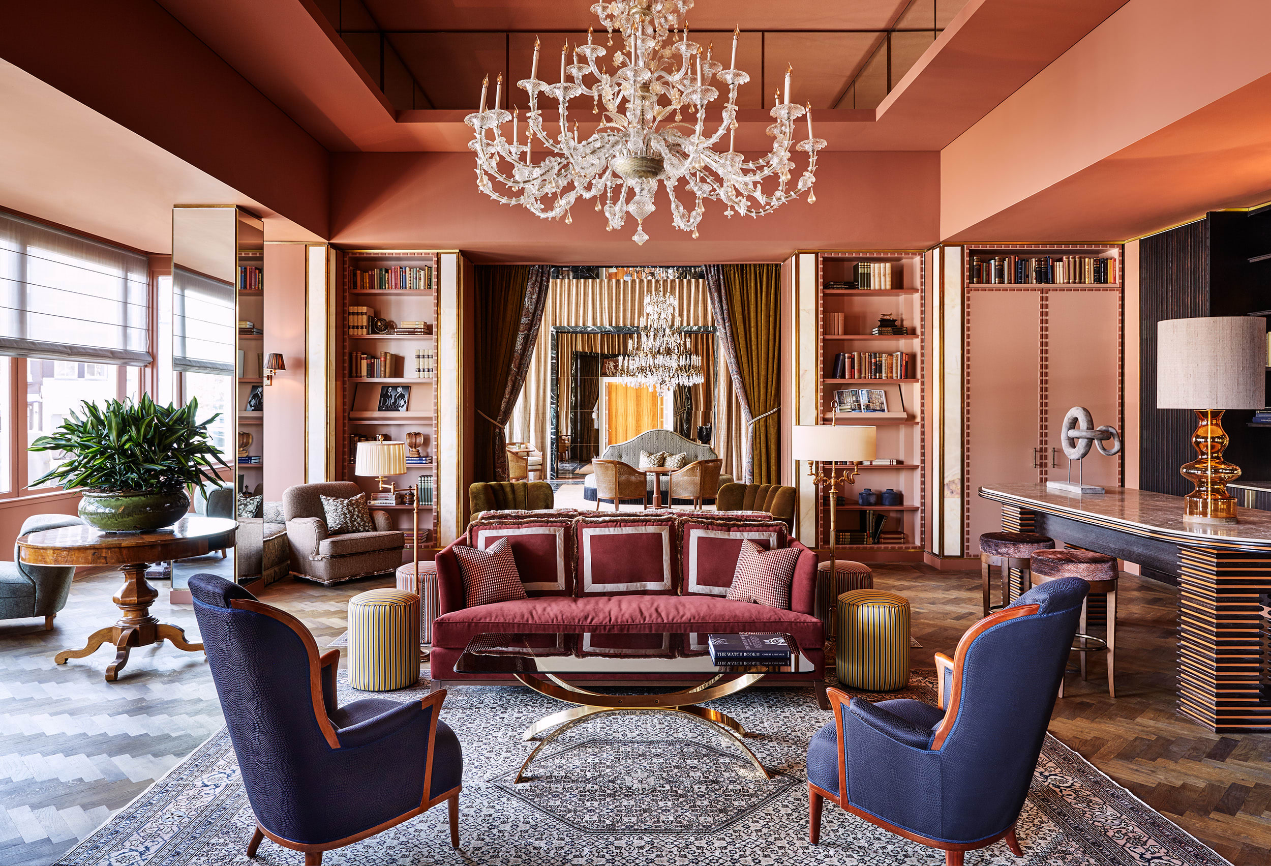 Elegant hotel lounge with blush-pink walls, a statement crystal chandelier, and plush velvet seating arranged around a polished coffee table, with built-in bookshelves and a sleek bar adding a refined, residential feel.