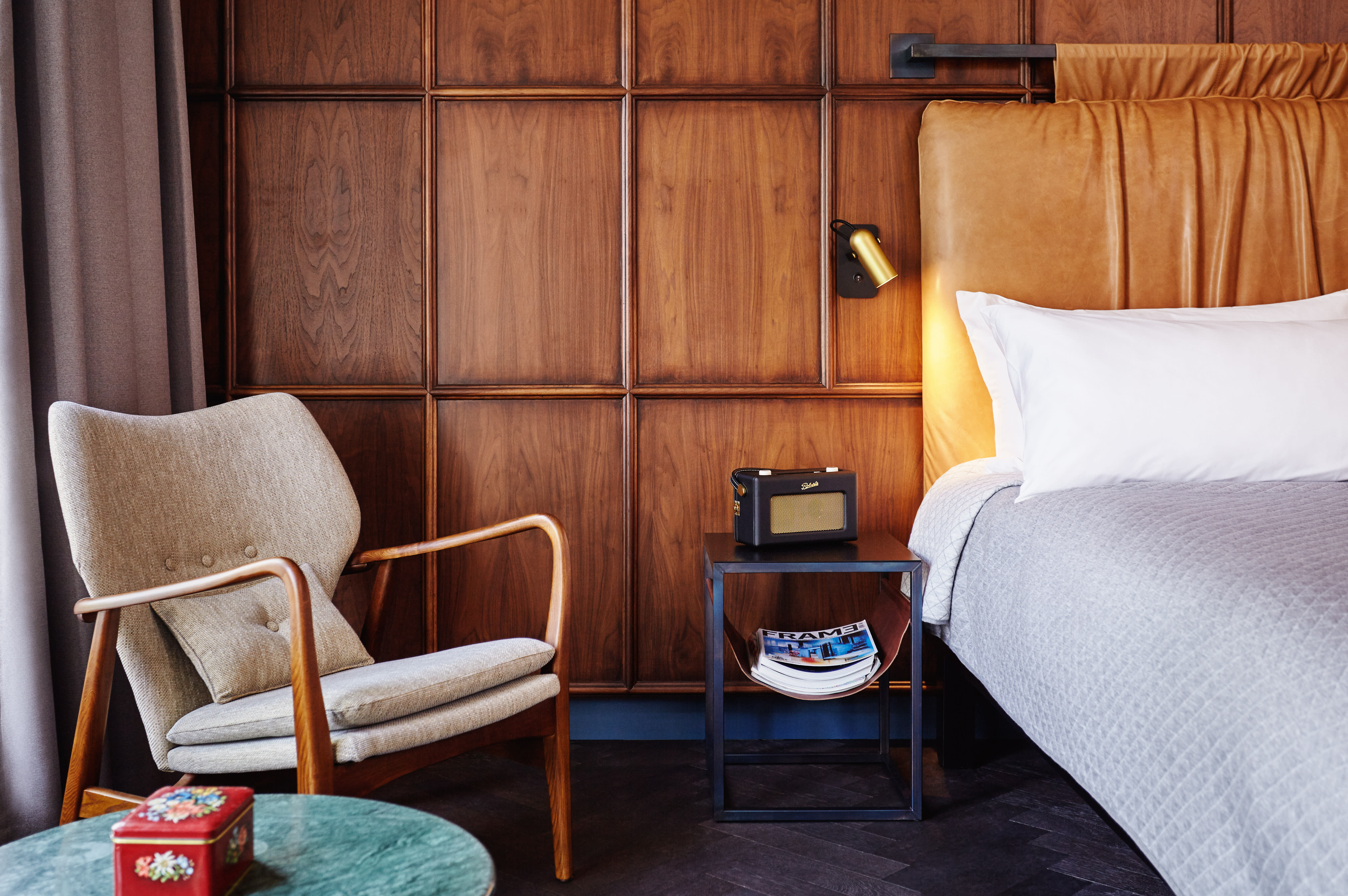 Warm, mid-century-inspired bedroom with a paneled wood wall, tan leather headboard, soft grey bedding, and a reading chair beside a small side table with books.