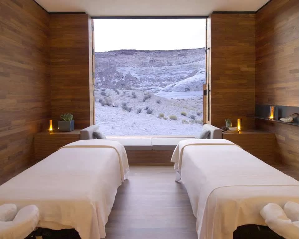 two side-by-side massage tables with white sheets in a wood room and hilly landscape outside
