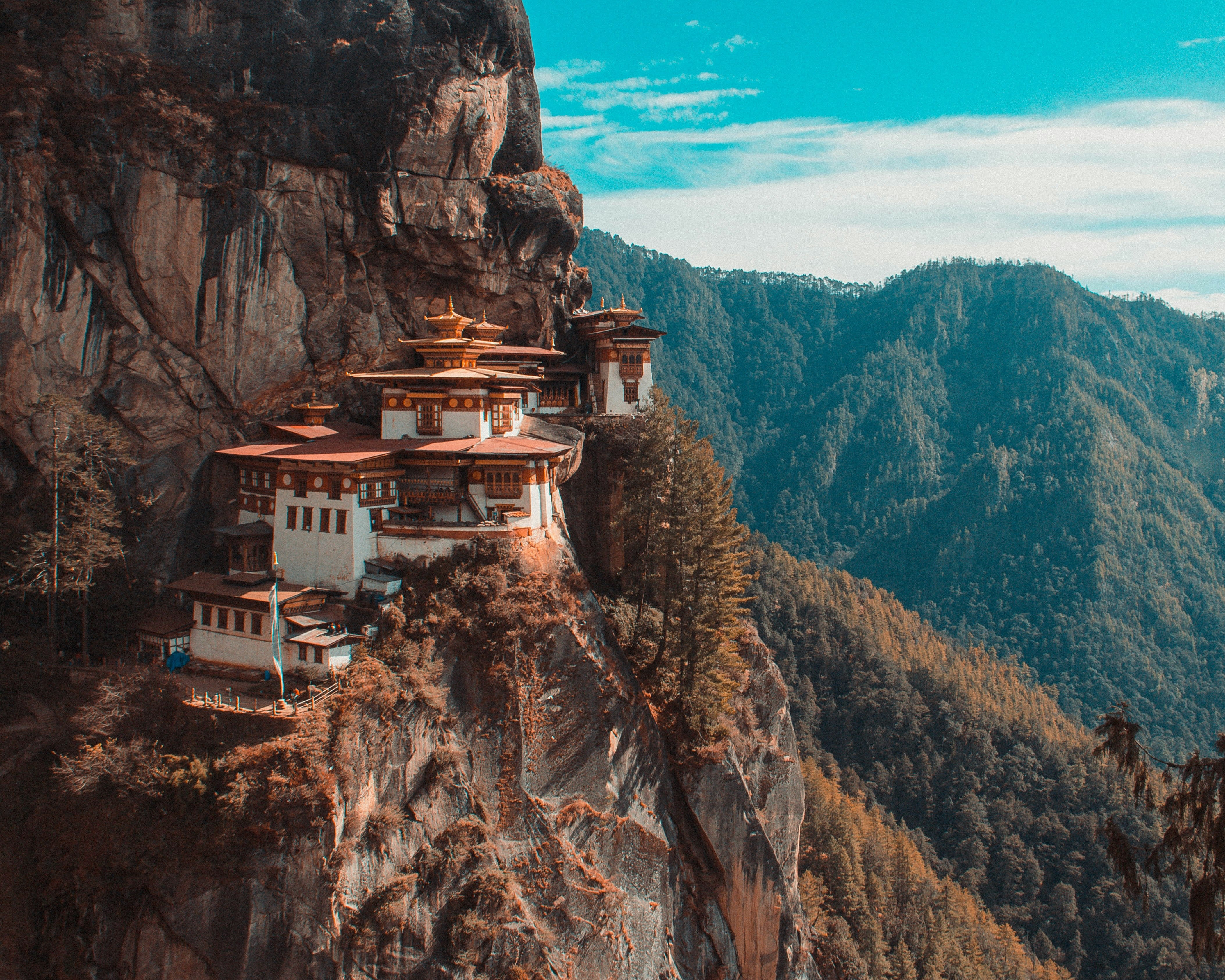 white and brown monastery on the edge of a cliff with tree covered mountains in the background during day