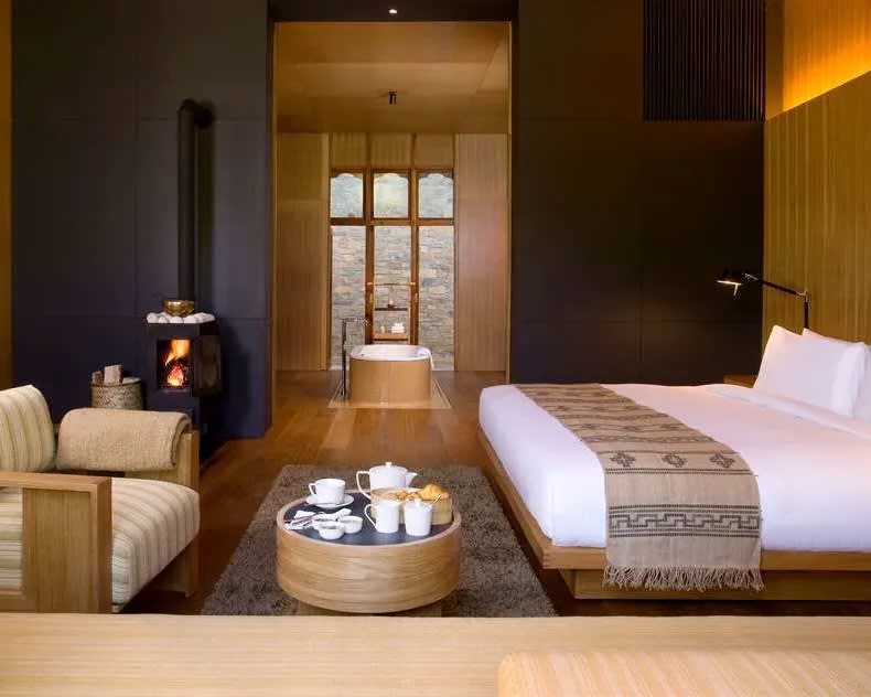 wood hotel room with white bed, tan cloth chair, and table with tea set during day