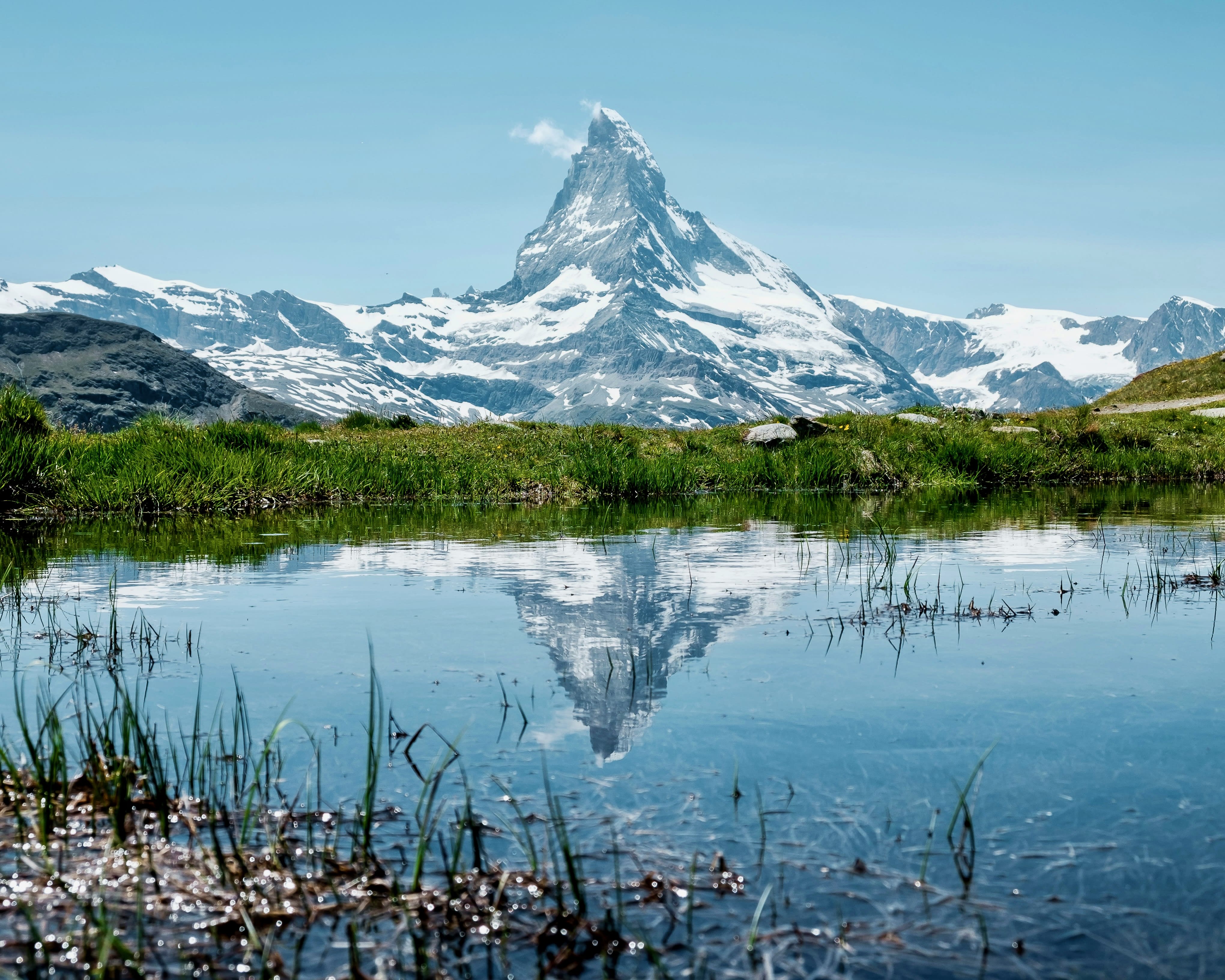rocky grey mountain peak with patches of snow reflecting in a green meadow lined lake