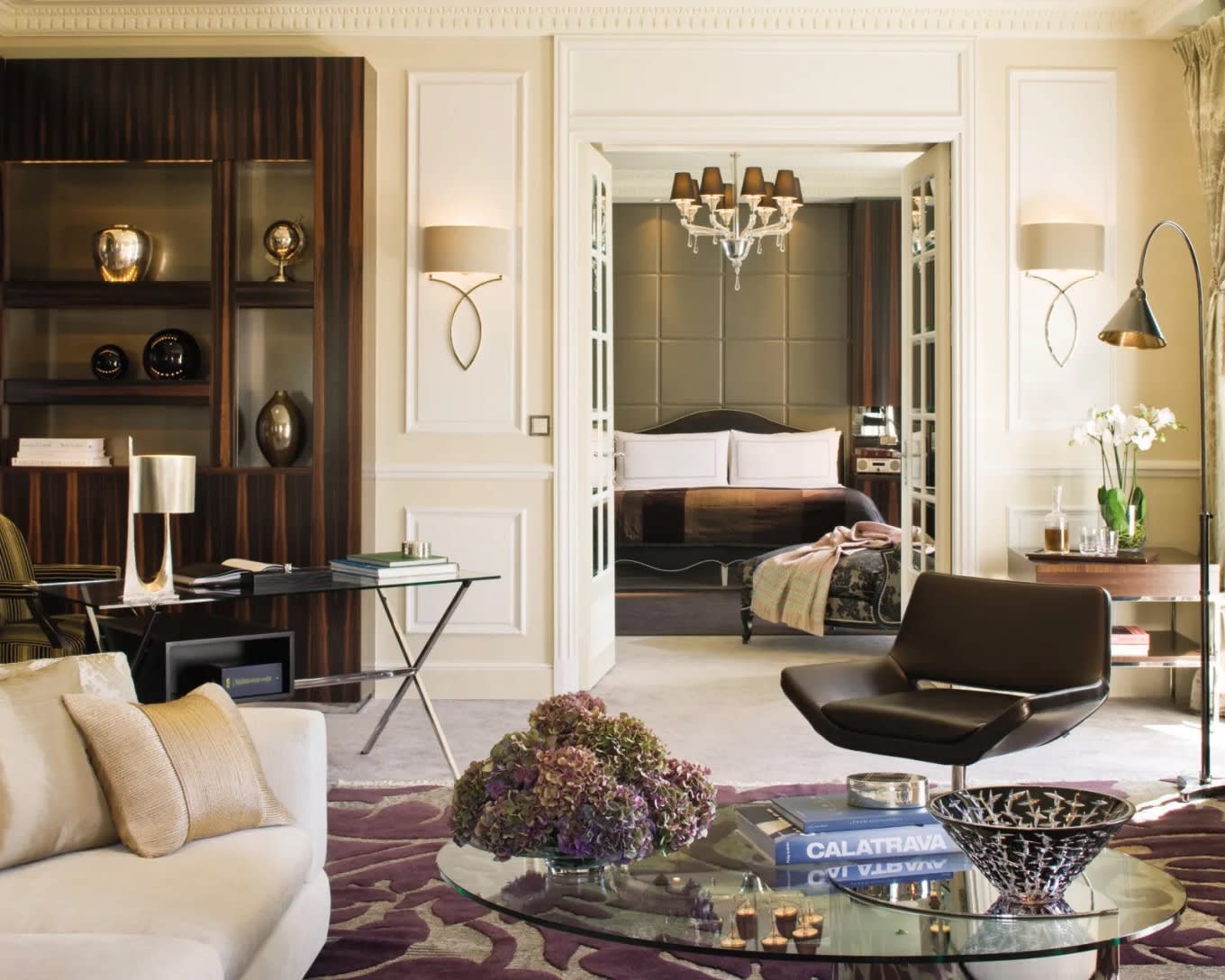 modern suite with white paneled walls, dark wood shelf, and living room furniture in front of open bedroom doors with bed under a chandelier in background