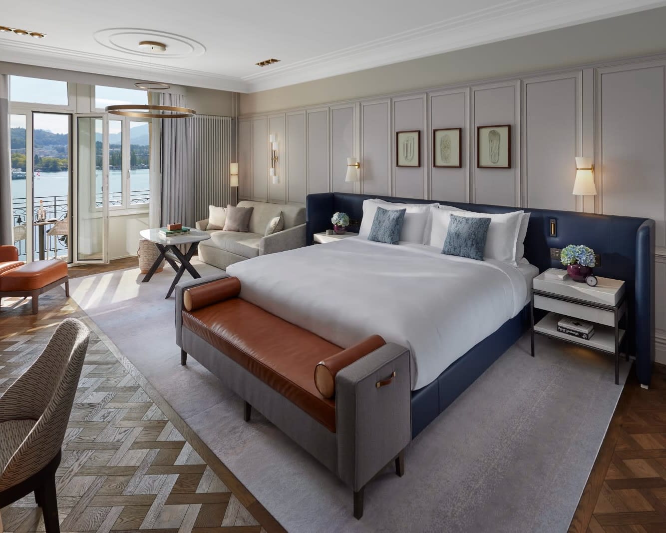 white bed with navy blue headboard, tan ottoman, and herringbone wood floor next to open windows