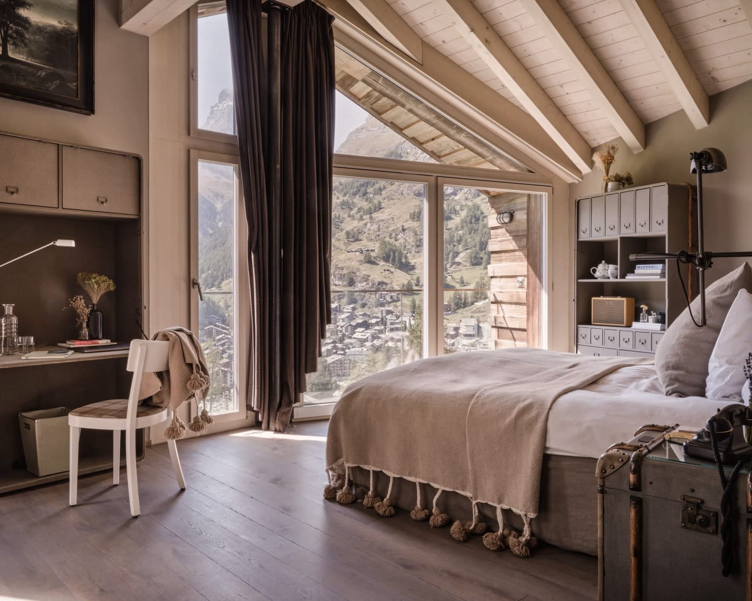 grey bed next to a white desk and chair with floor-to-ceiling windows overlooking green mountains outside