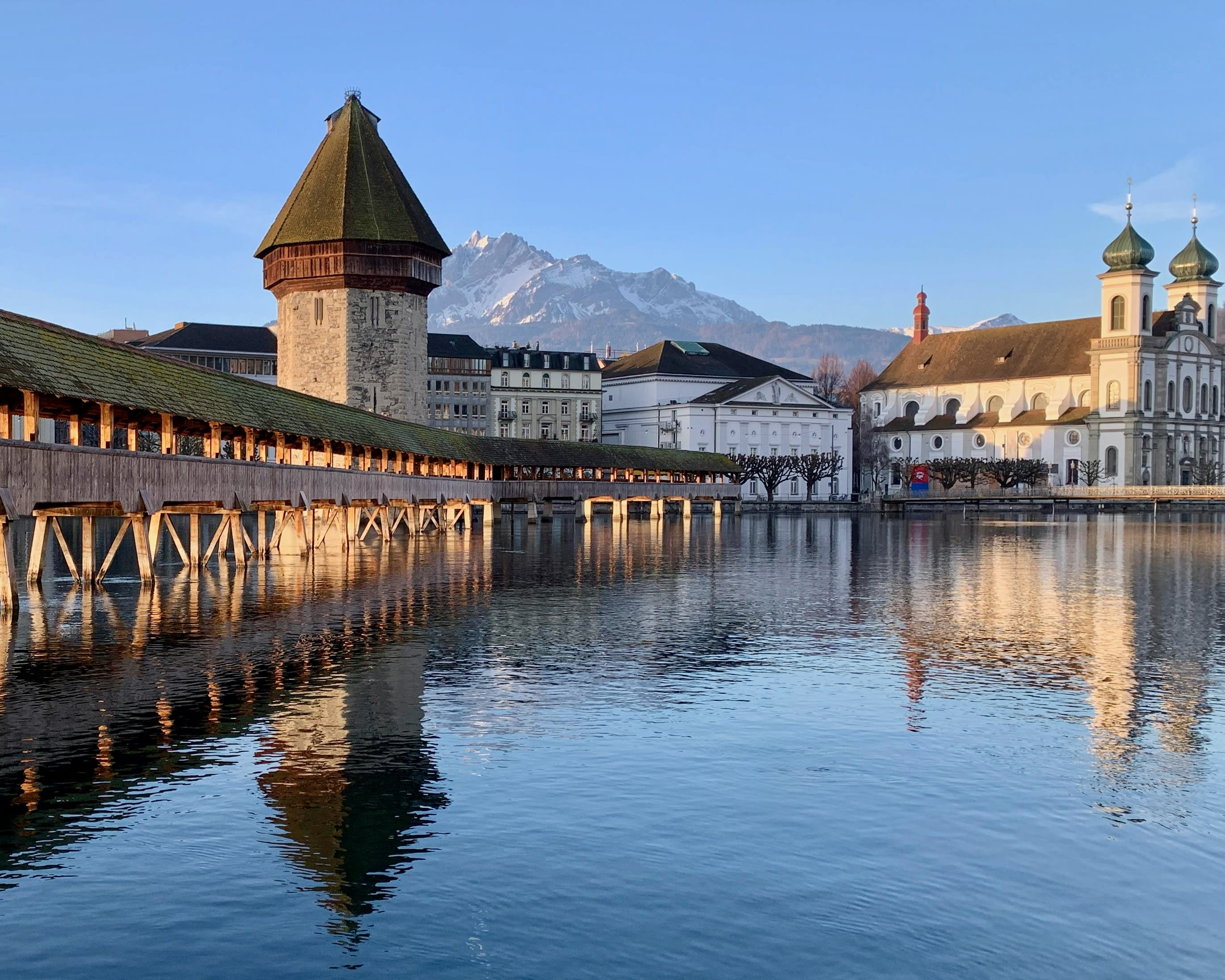 old wood bridge over a river with historic buildings and towers on the town behind with snow capped mountains in the background