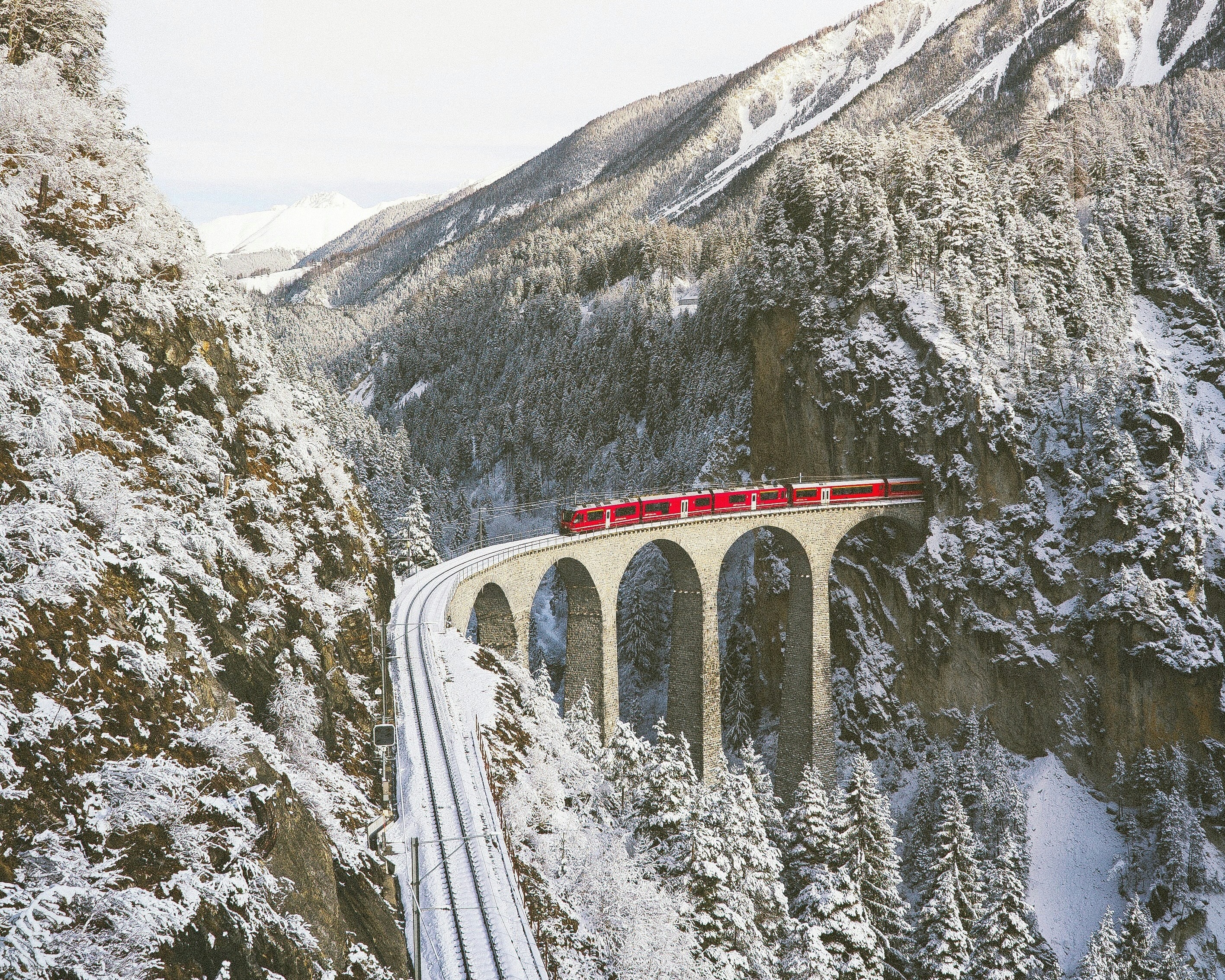 red train on stone bridge over a snowy mountain gorge with trees surrounding the tracks during day