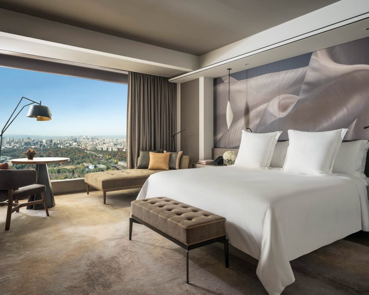 white hotel bed with grey ottoman and chaise lounge in front of textured modern grey wall and windows with green park and cityscape outside