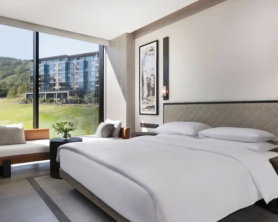 white bed next to windows overlooking green hills and modern black resort building