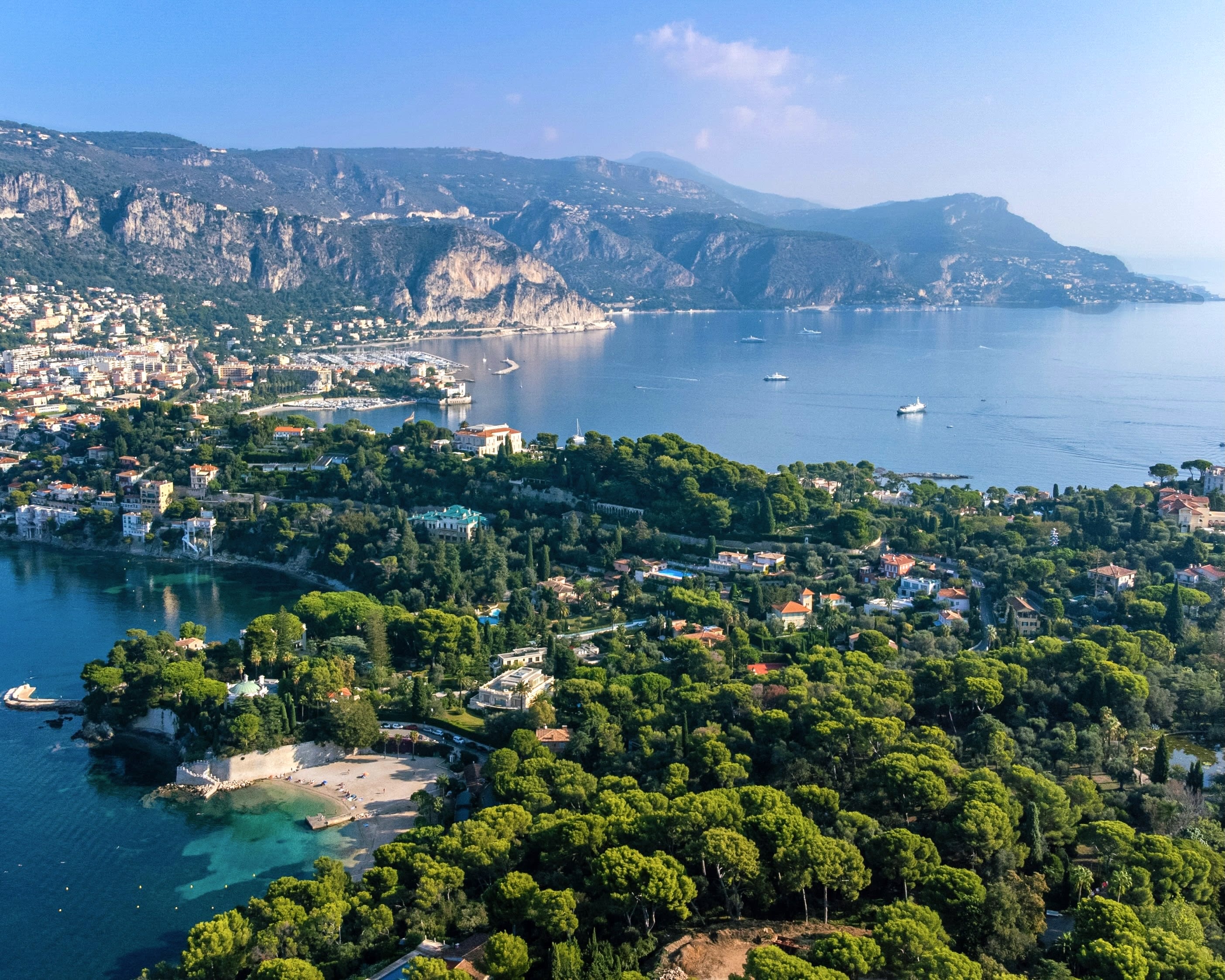 aerial view of green peninsula with rocky mountains in background surrounded by villas and turquoise water