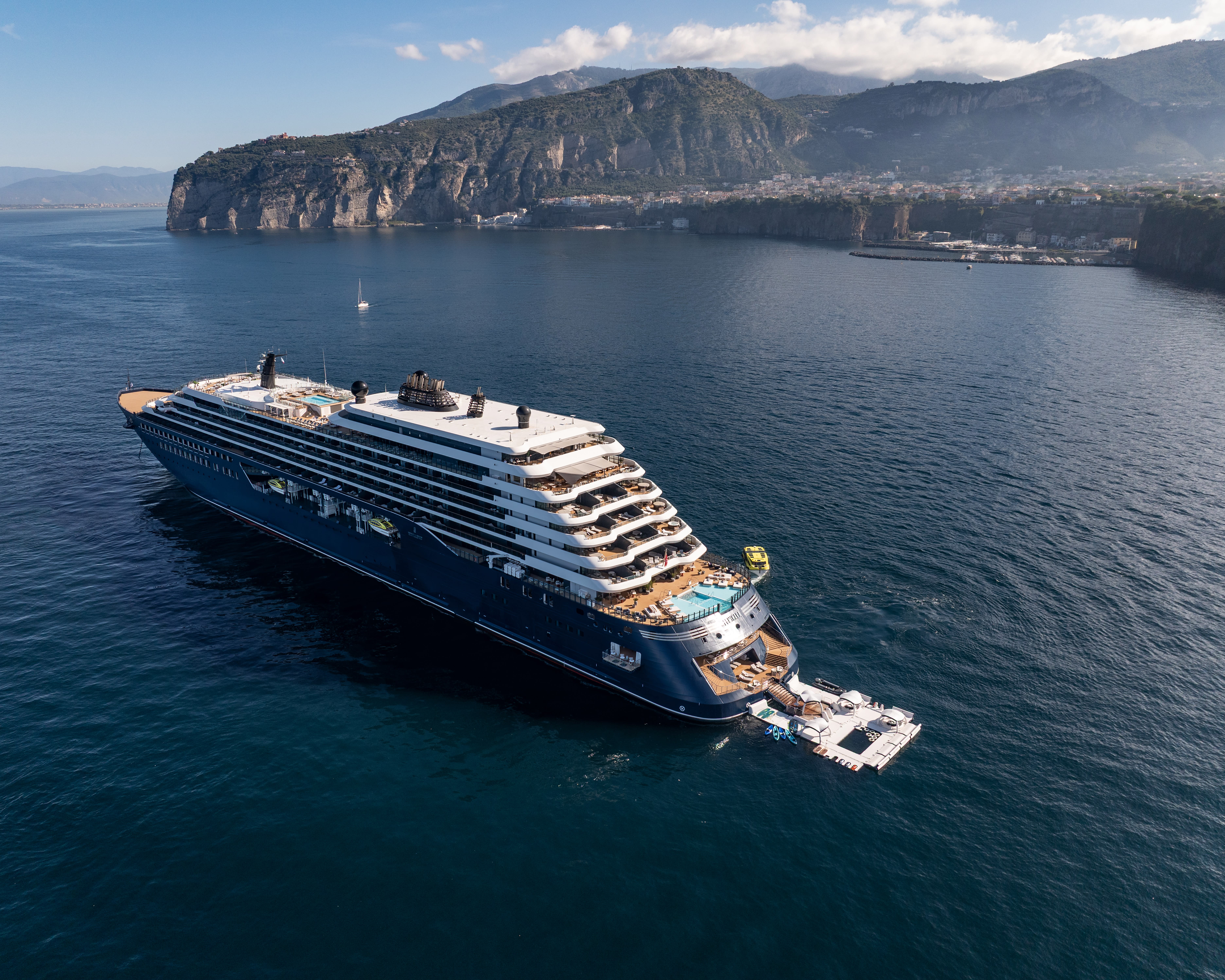 a large blue and white yacht anchored in blue ocean waters off a rocky mountainside town