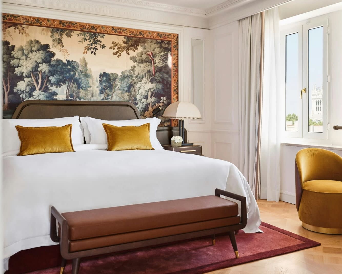 a white bed with maroon ottoman, gold pillows and side chair, and ornate drawing above the bed