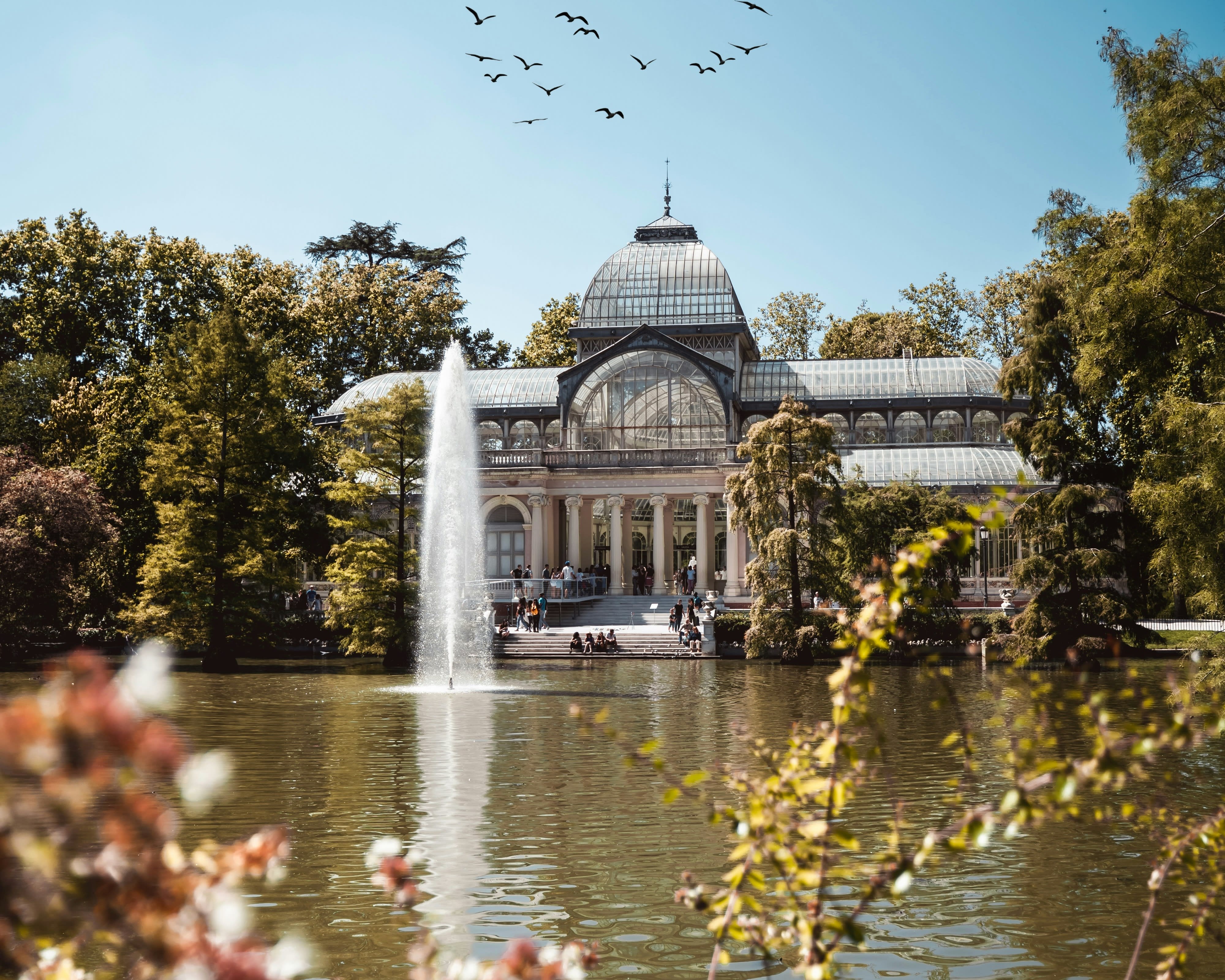 a glass botonical green house next to a lake with fountain and green trees around