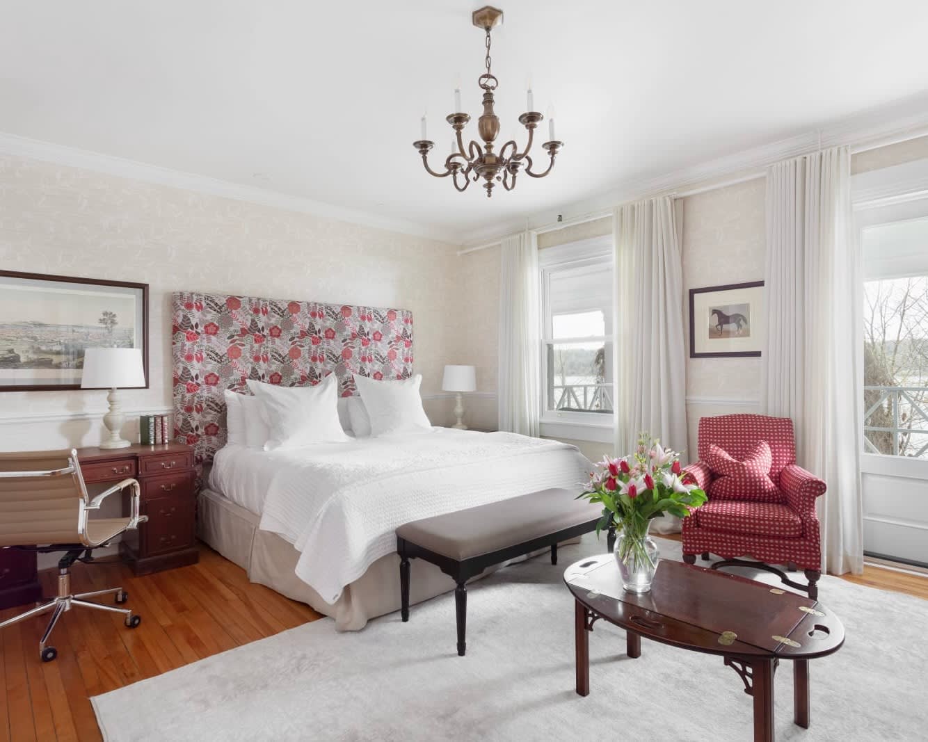 white hotel room with chandelier above a classical style white bed, with dark wood furniture and red accent chair with red headboard