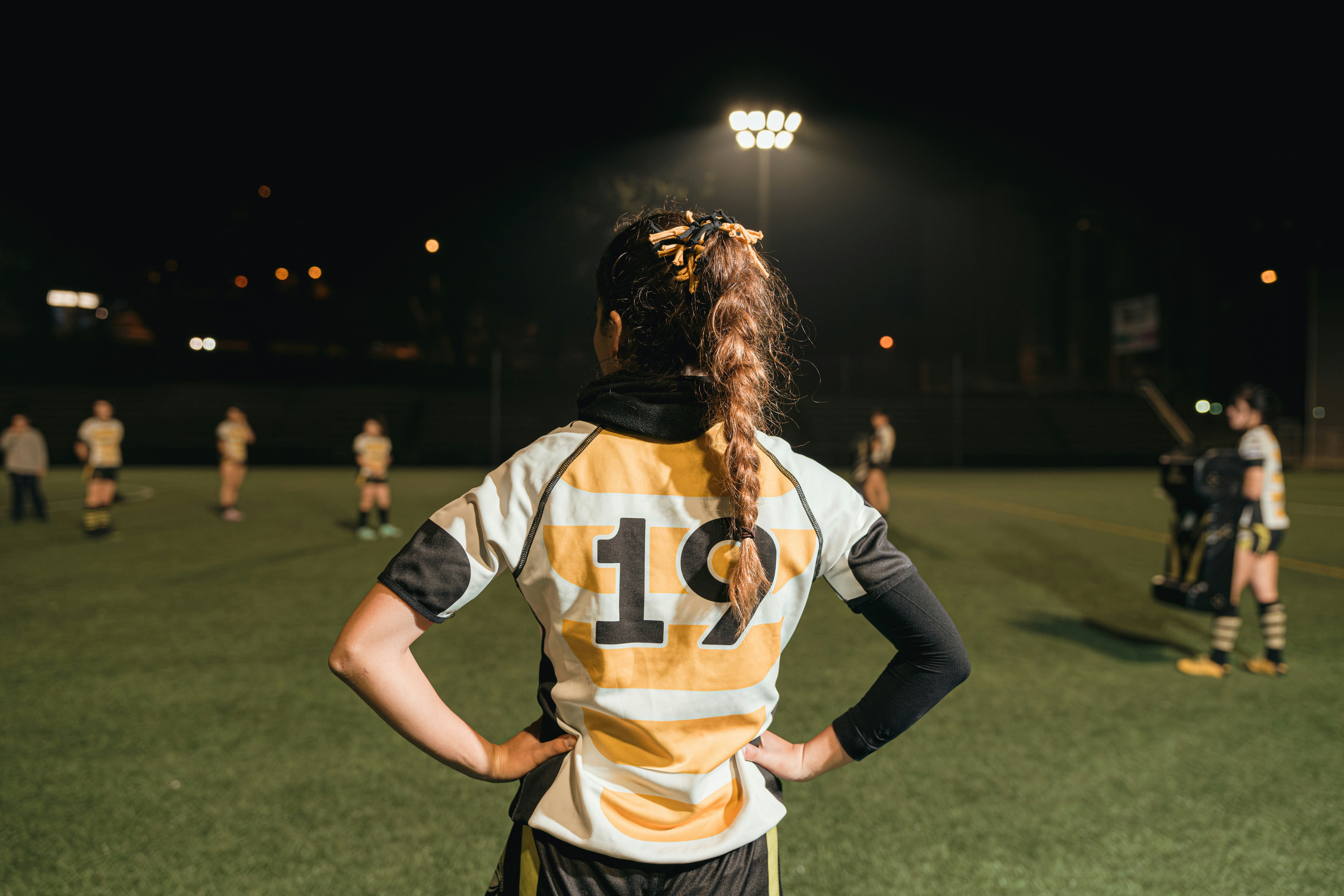 A girl in a yellow and white striped jersey stands on a rugby pitch with her hands on her hips