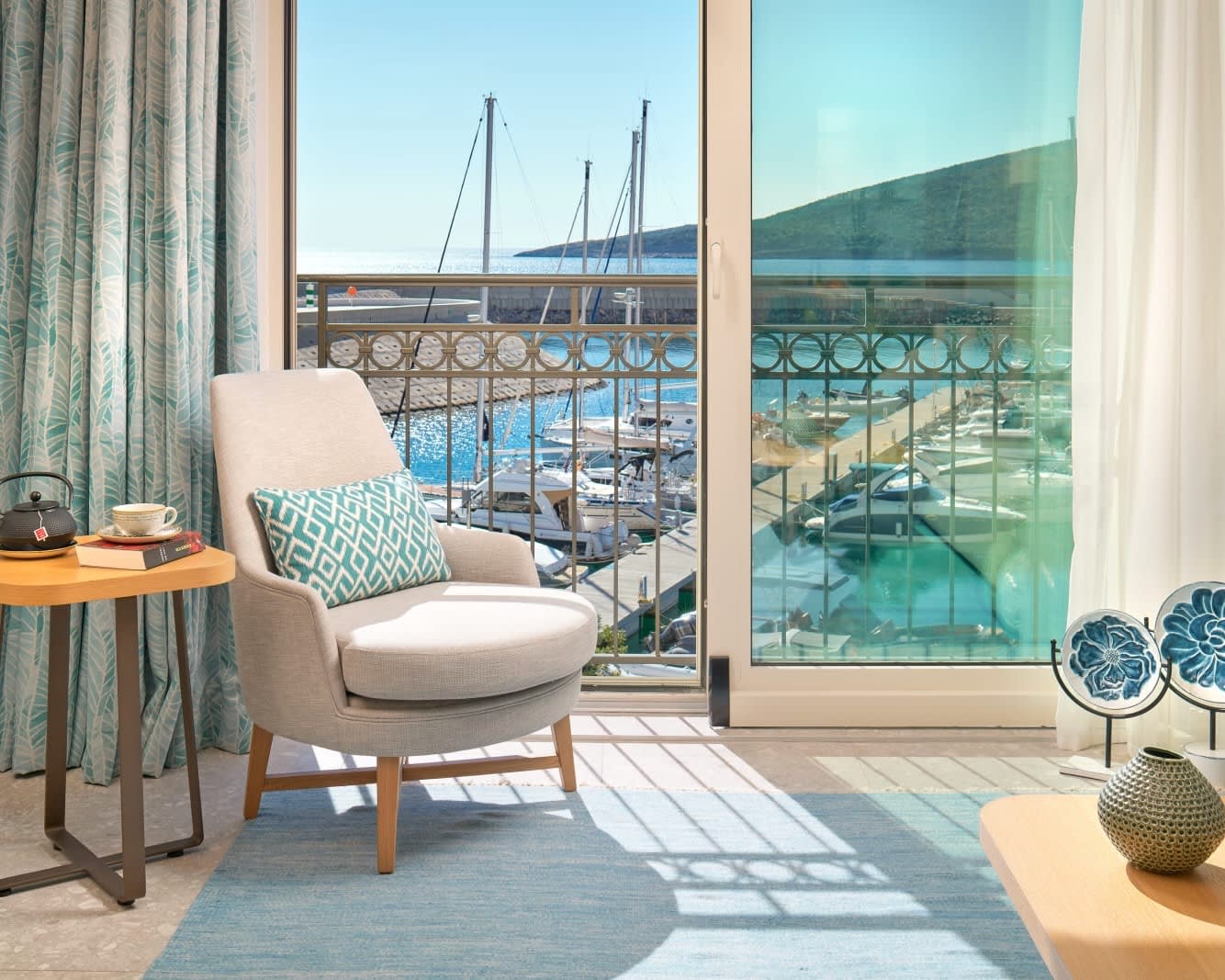 a cream colored chair with turquoise pillow in front of sliding glass doors with yacht marina outside during day