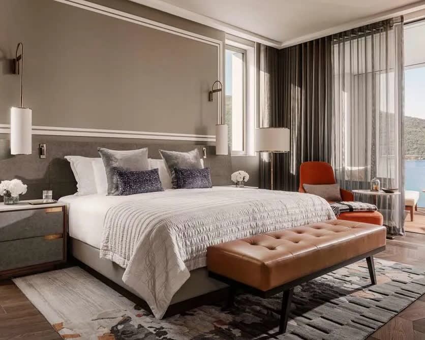 Modern room with white covered bed, brown ottoman at foot of bed, and grey walls behind the bed.