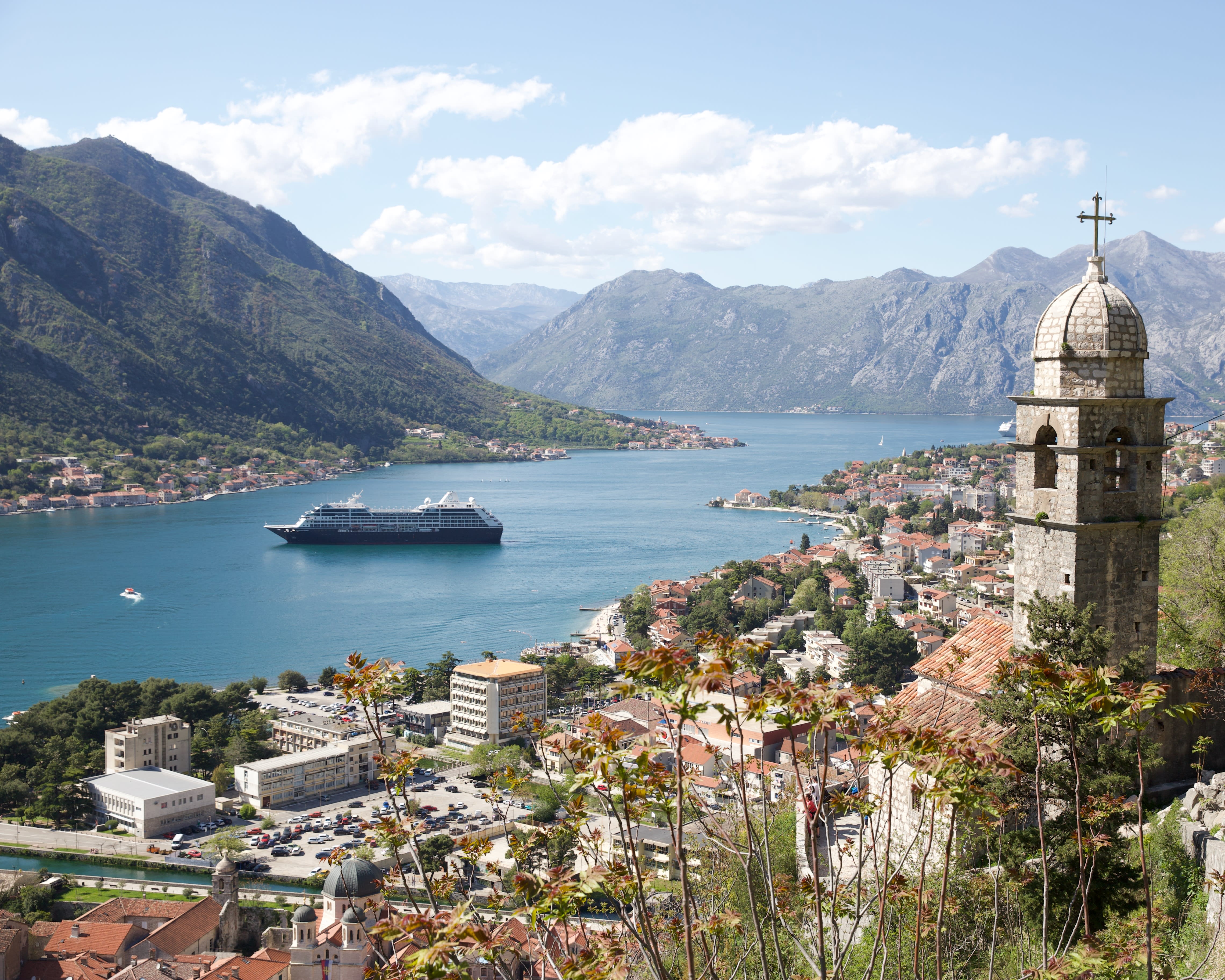cruise ship in the middle of a blue bay with ancient tower in foreground and buildings at the foot of hills around the bay