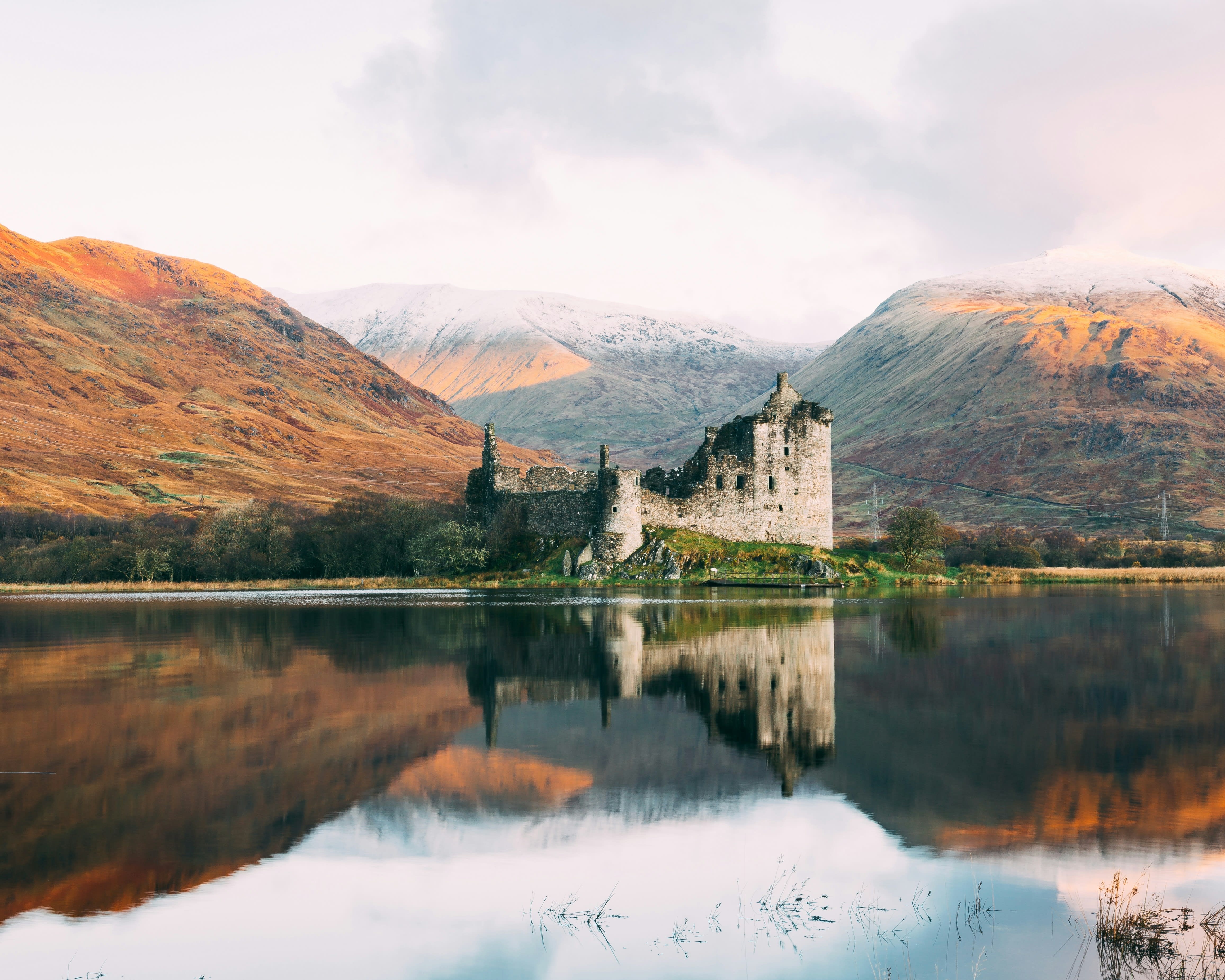 ruined castle reflected in the lake with tan hills in the background under cloudy sky