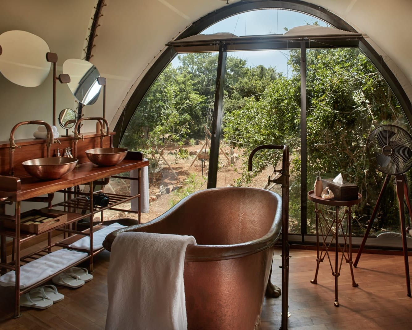 A copper freestanding bathtub in the middle of a tented bathroom with jungle outside