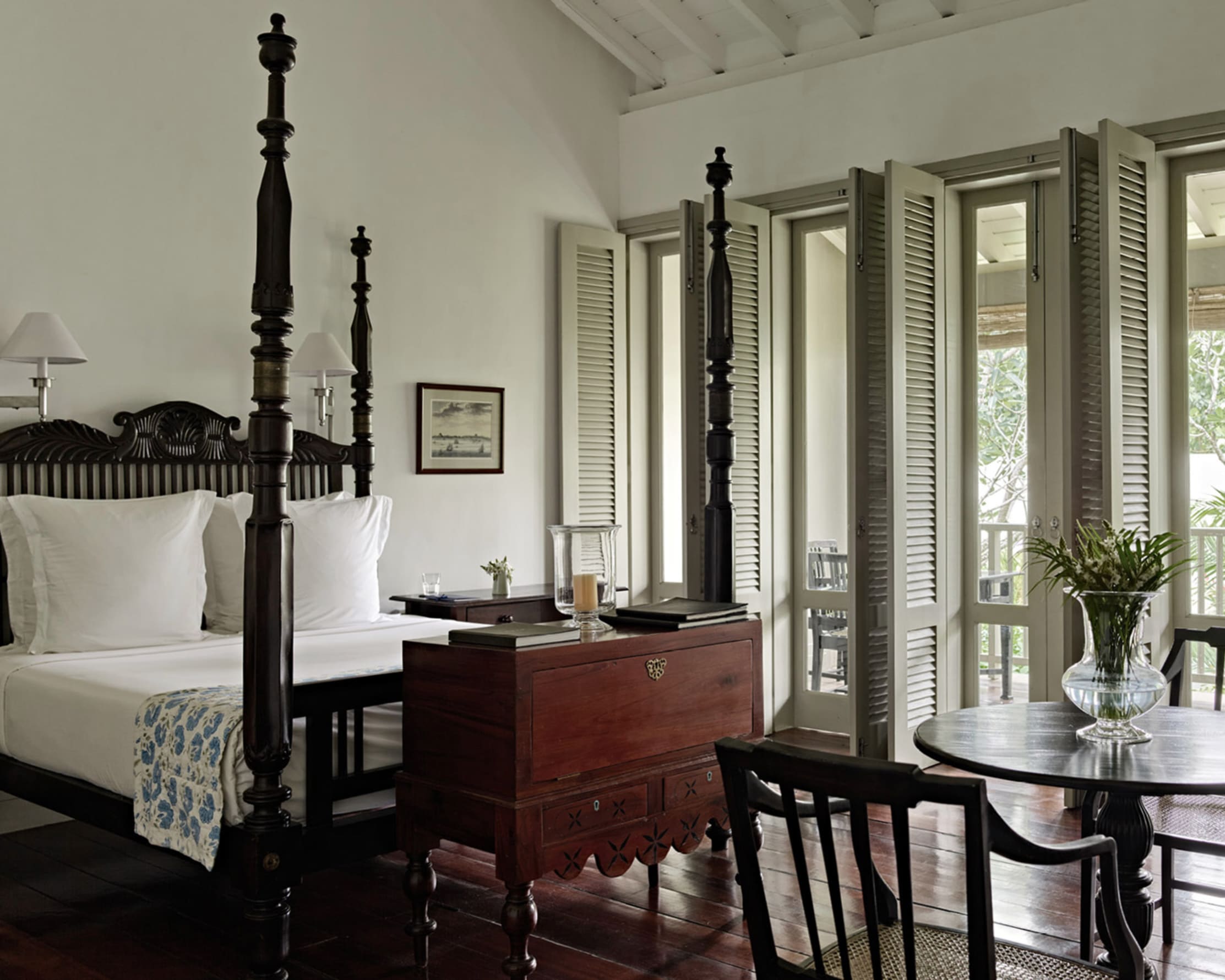 Black four poster bed with white linens, wood cabinet at the foot of the bed, and open shutters leading outside