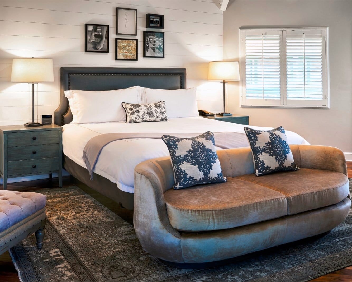 A blue wood bed with brown love seat at the foot of the bed and framed art above the bed