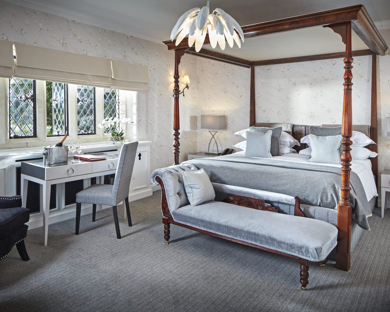 wood four poster bed with grey bed spread, grey bench at the foot of the bed, and grey carpets, with white desk under windows