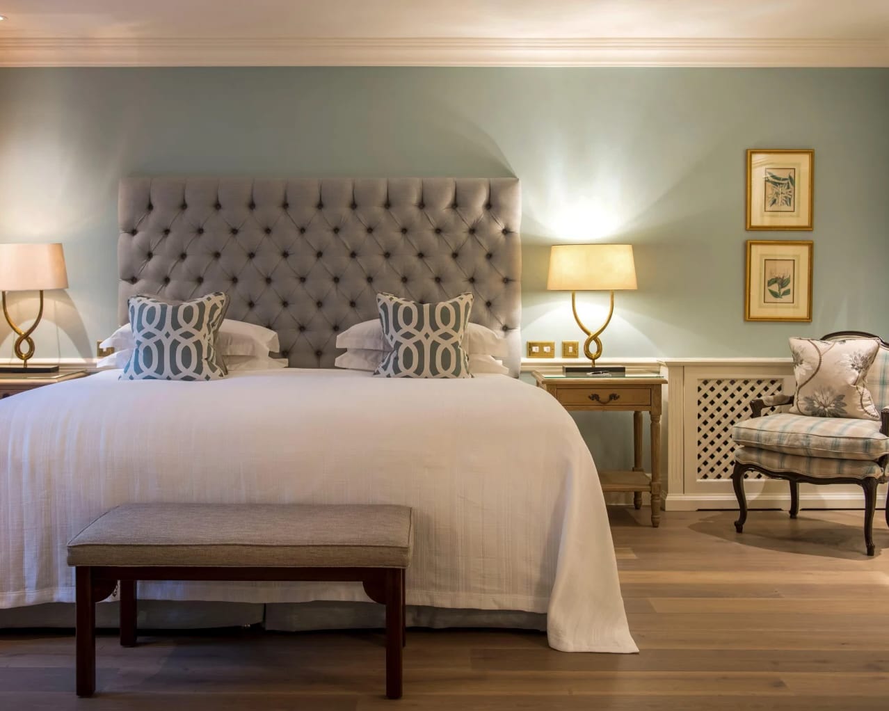 A bed with white bedspread, and grey padded headboard with traditional chair next to it