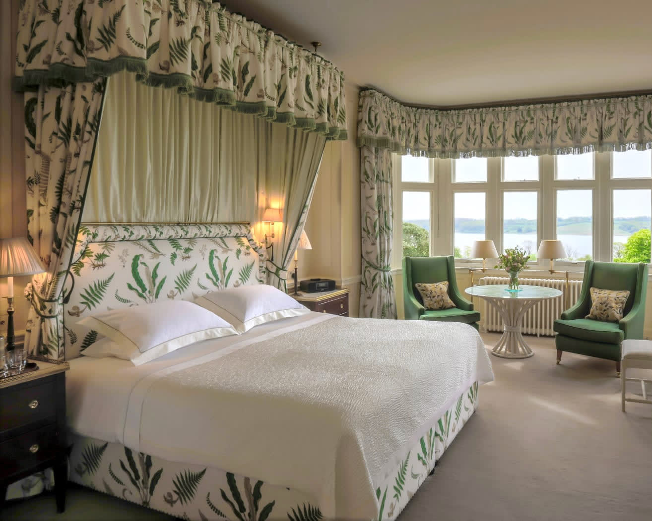 white bed with white and green leaf pattern fabric headboard, drapes over bed, and curtains with green chairs