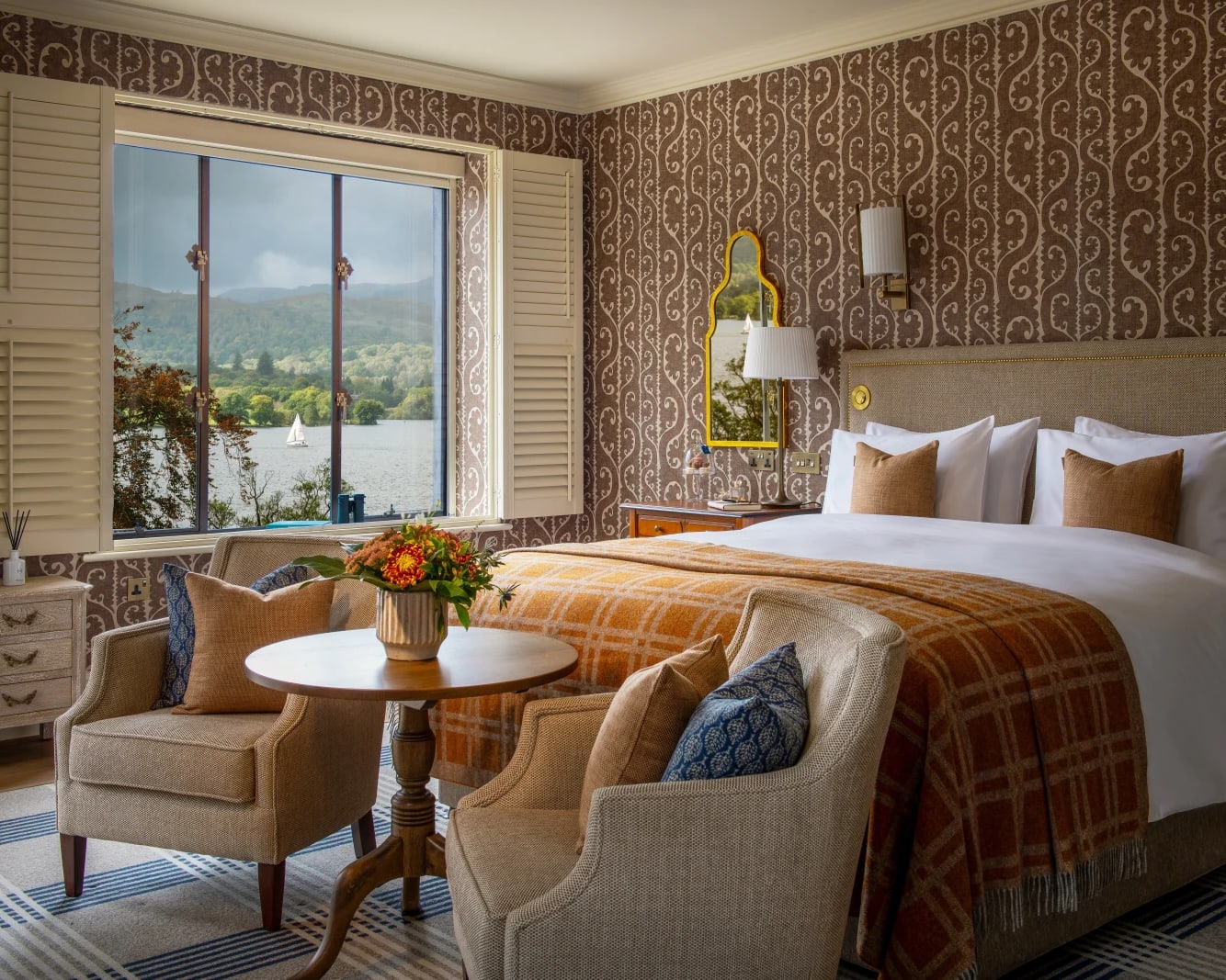 brown patterned wall paper bedroom with white bed, orange blanket, and tan chairs