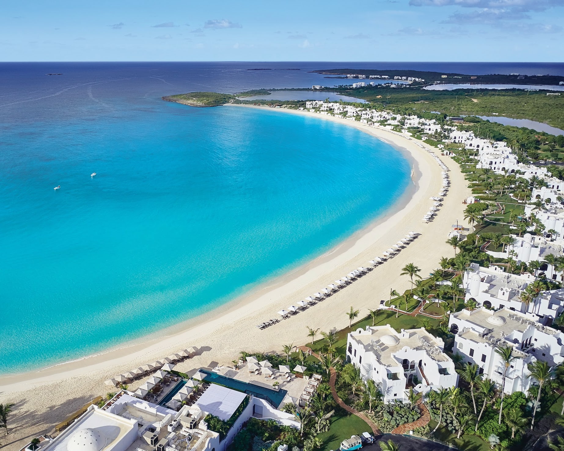 Aerial view of a crescent white sand beach with white villas among green palm trees and turquoise waters off shore