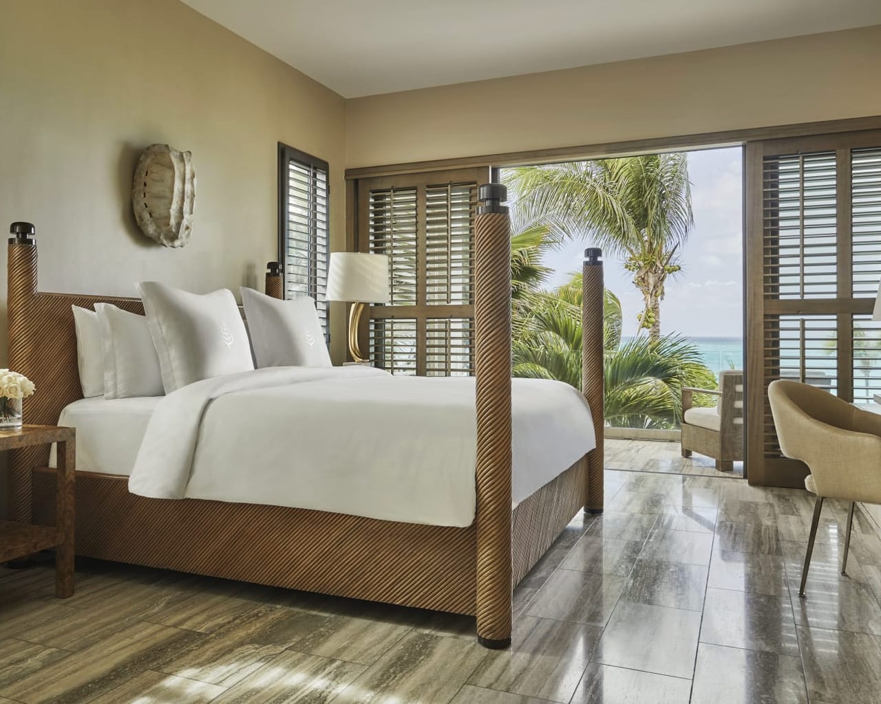 Hotel room with brown four poster bed, white linens, and open shutters onto tropical beach with palm trees
