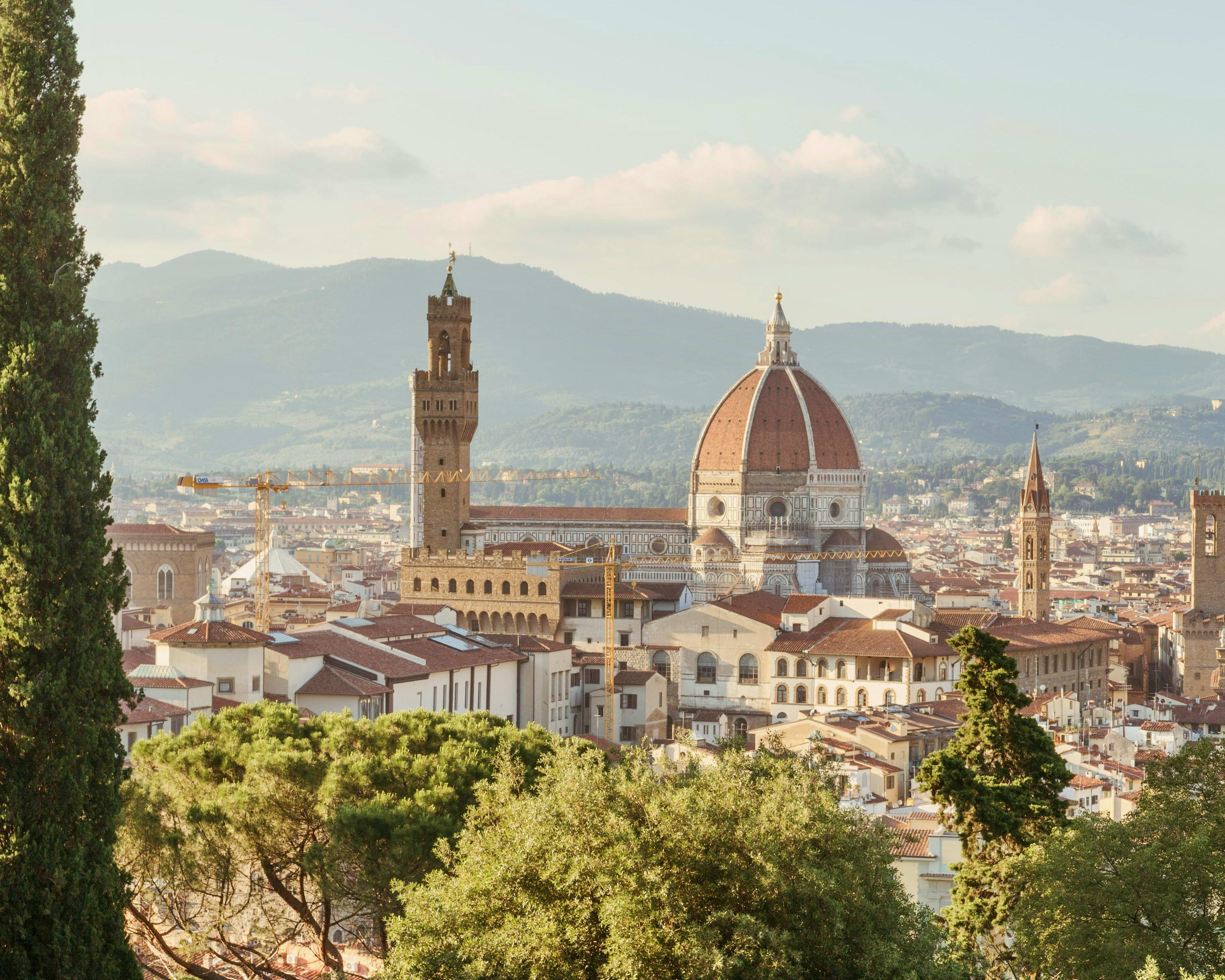 red rooftops across a city with a tower and red dome cathedral in the center, trees in the foreground, and mountains in the distance