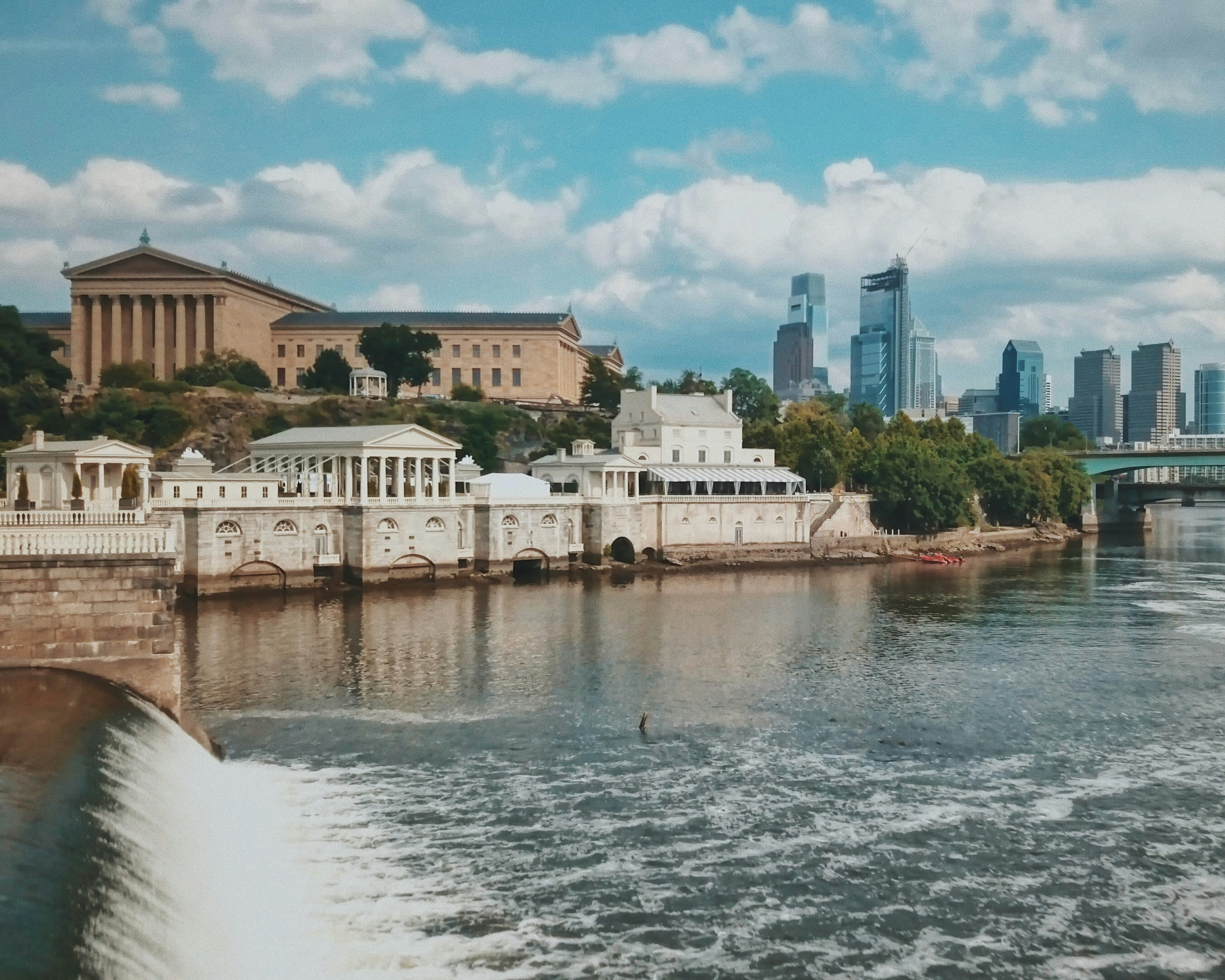 a waterfall and river in front of white and tan columned buildings along the water and skyscrapers in the background