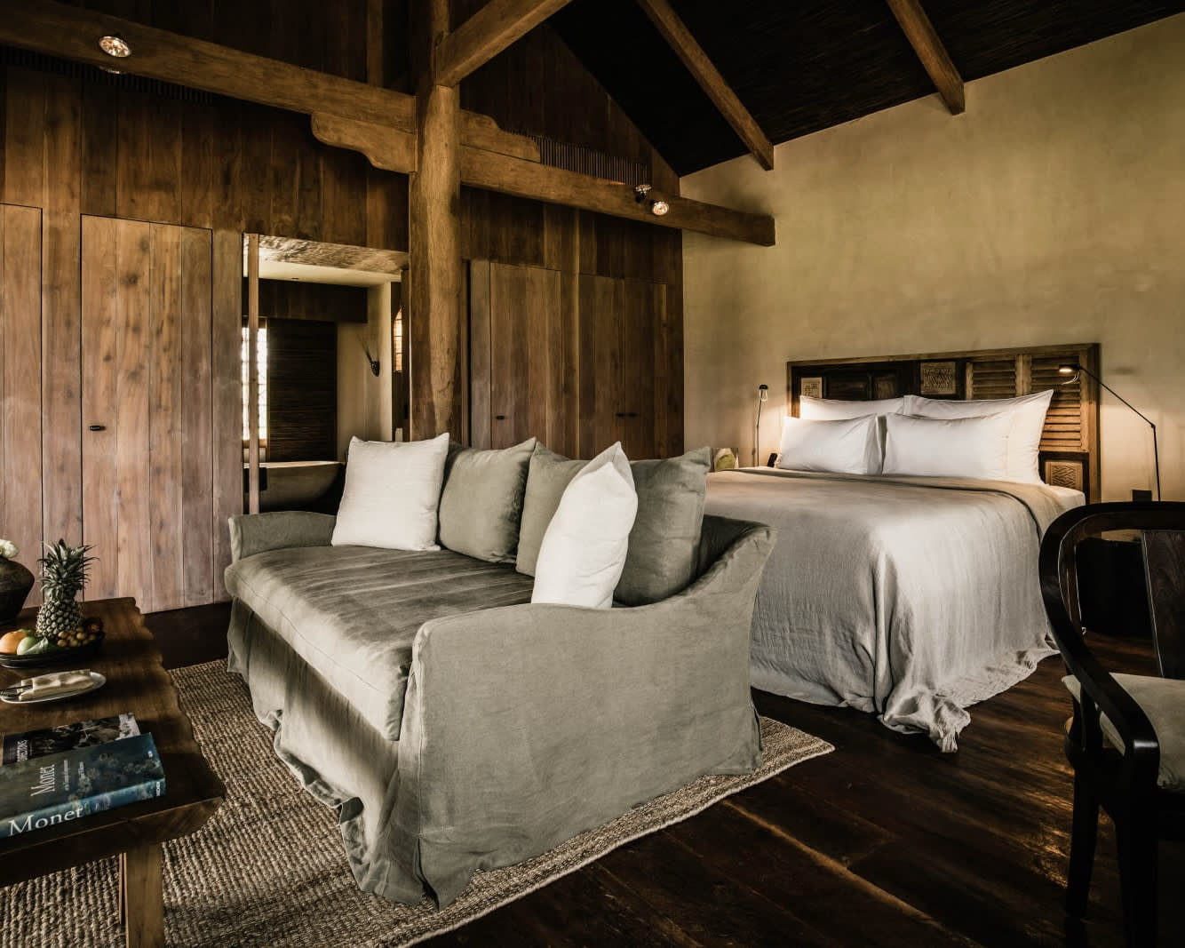 white sheet covered bed with a grey sofa at the foot of the bed in a wood walled and roofed room