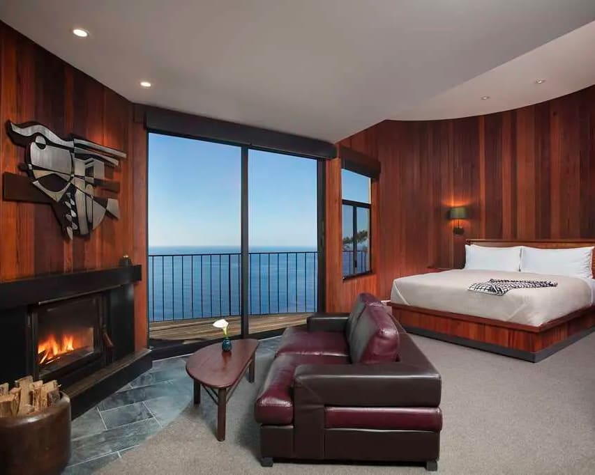 wood walled hotel room overlooking the ocean with fireplace, brown leather chair, and white sheet covered bed