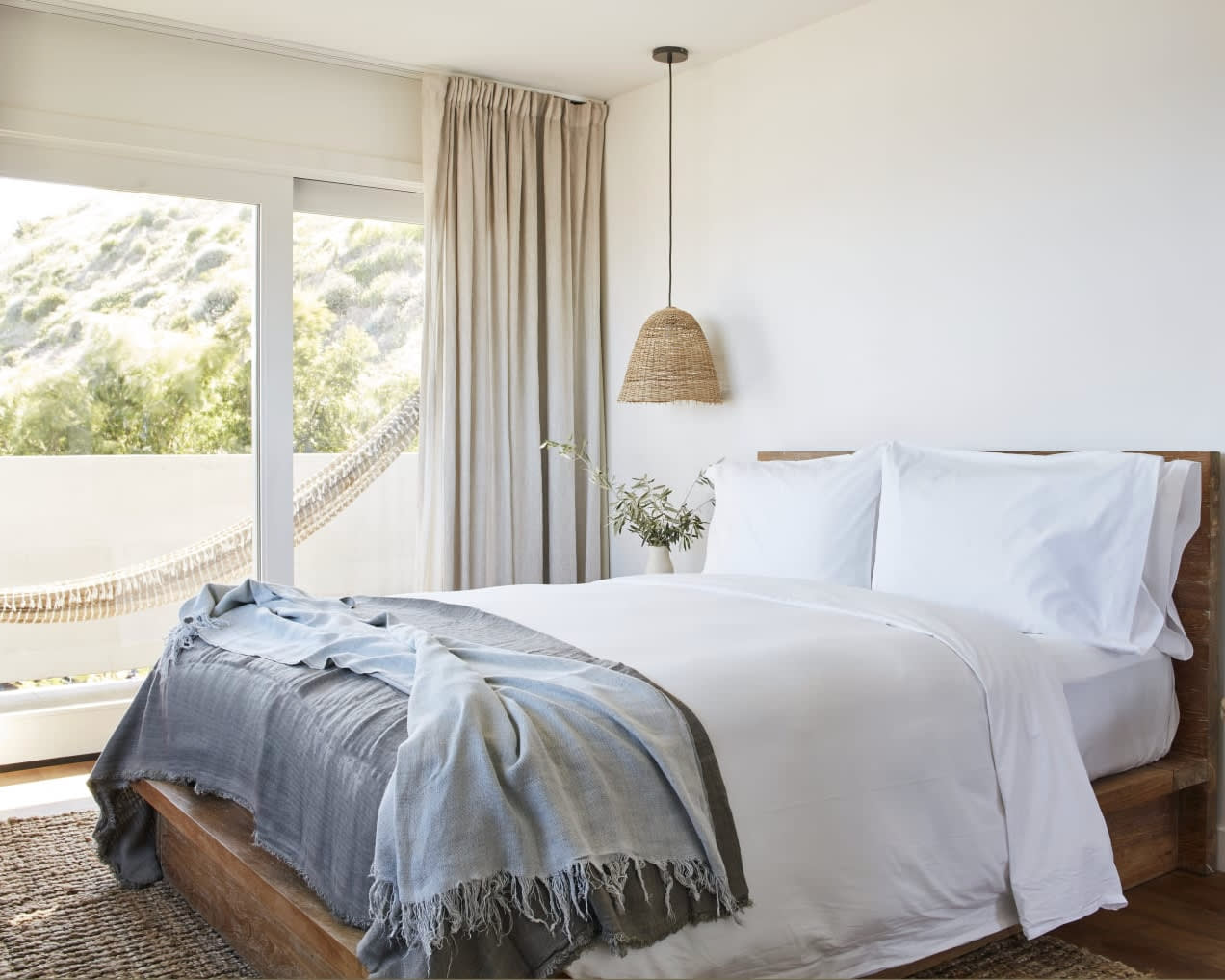 modern white hotel room bed with blue blanket, wood frame, and tan hammock on the balcony outside