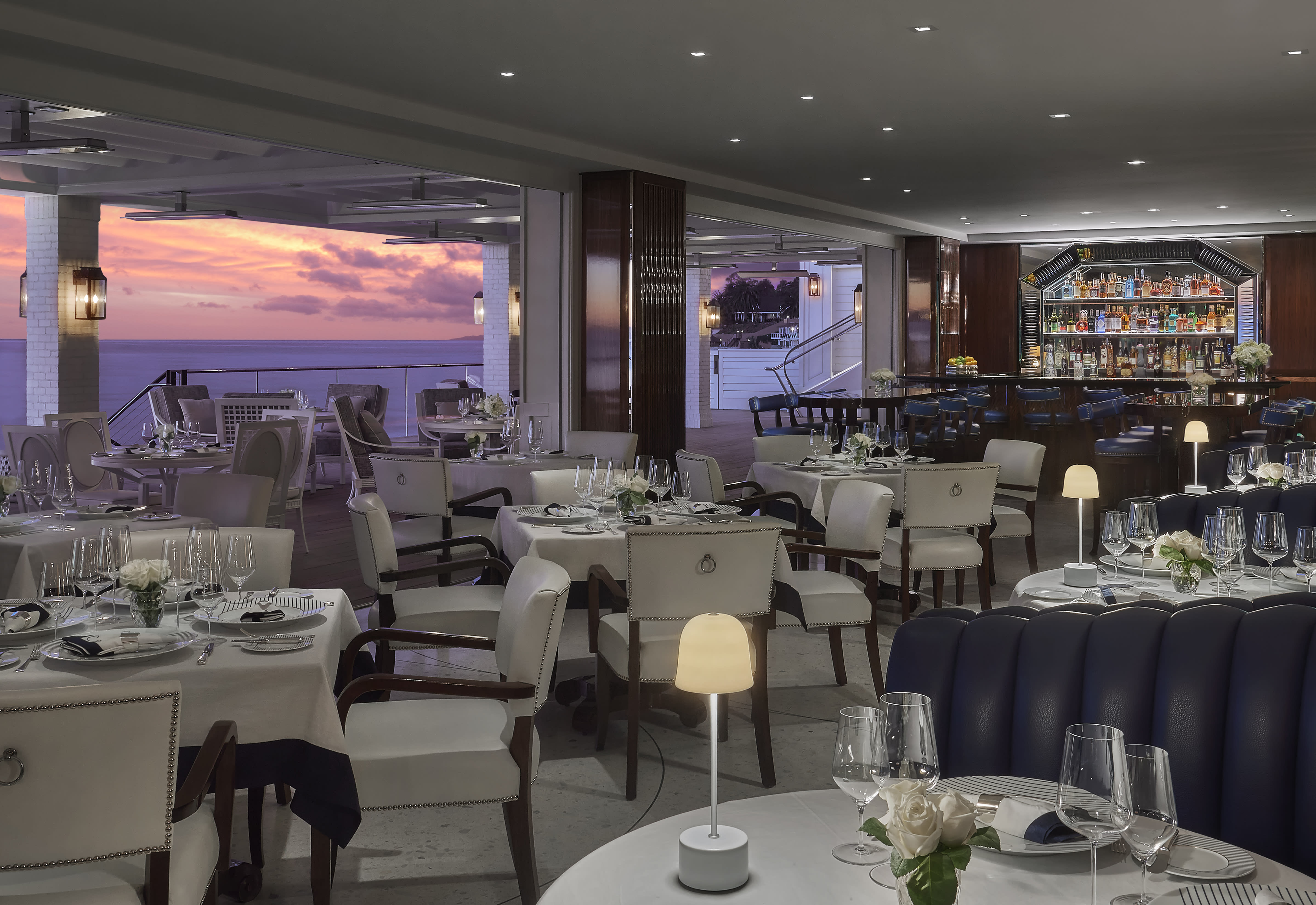 a fine dining restaurant with white and black chairs, tabletop lights, and a sunset over the ocean outside