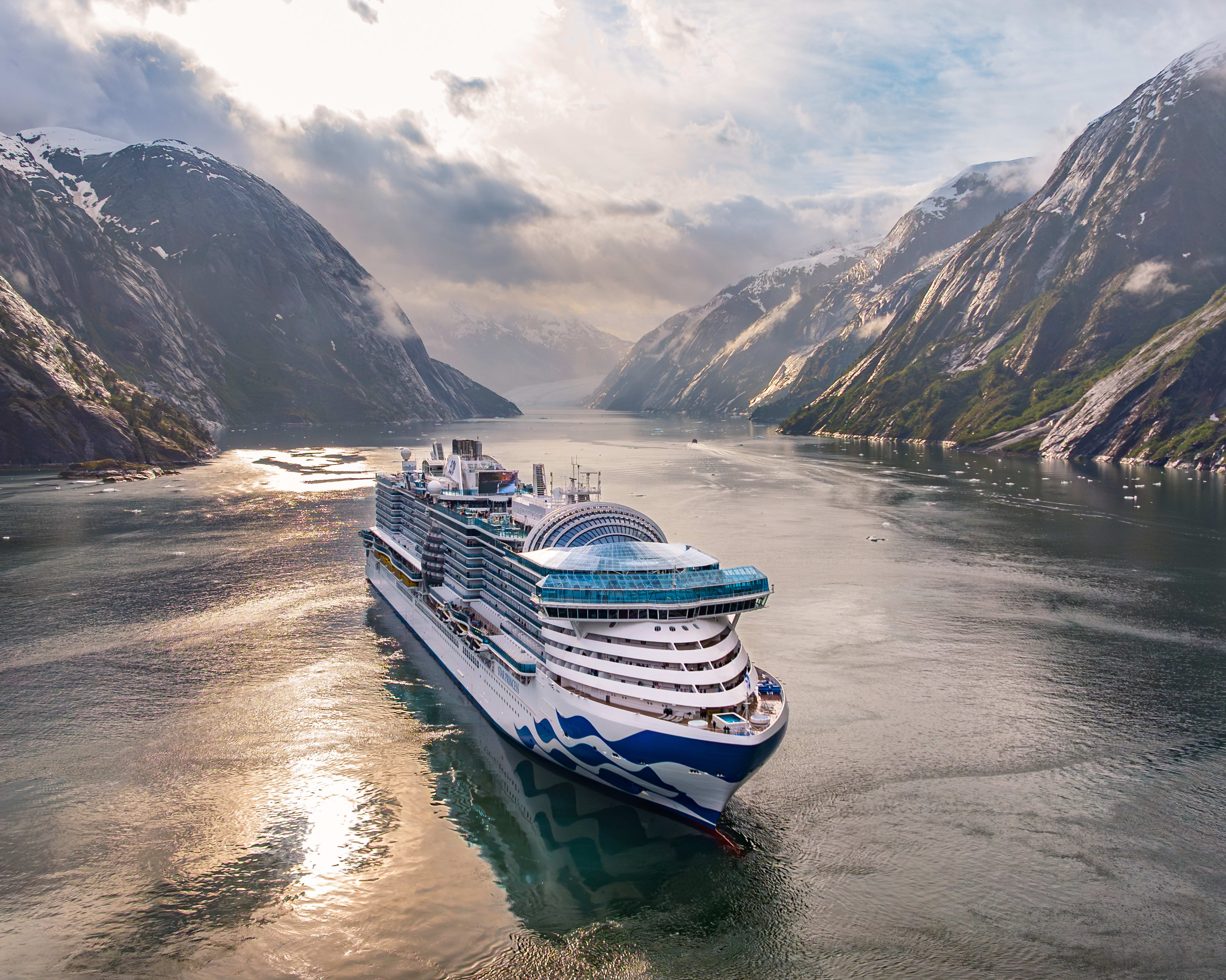 Large modern white and blue cruise ship sailing in a fjord with snowy rocky mountains on both sides during day