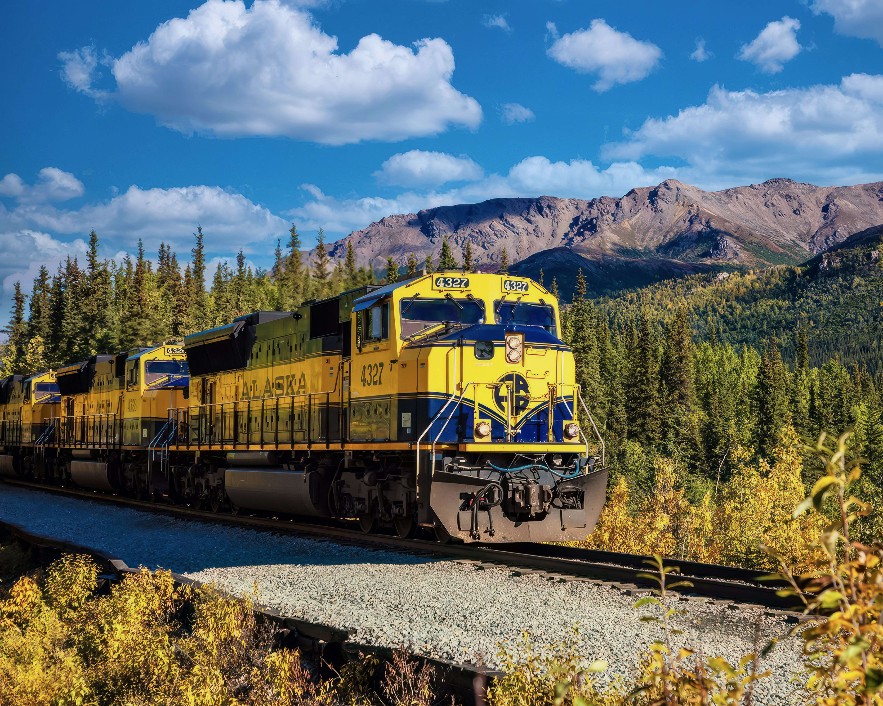 bright yellow and navy blue train in remote wilderness with green trees, brown mountains, and blue sky