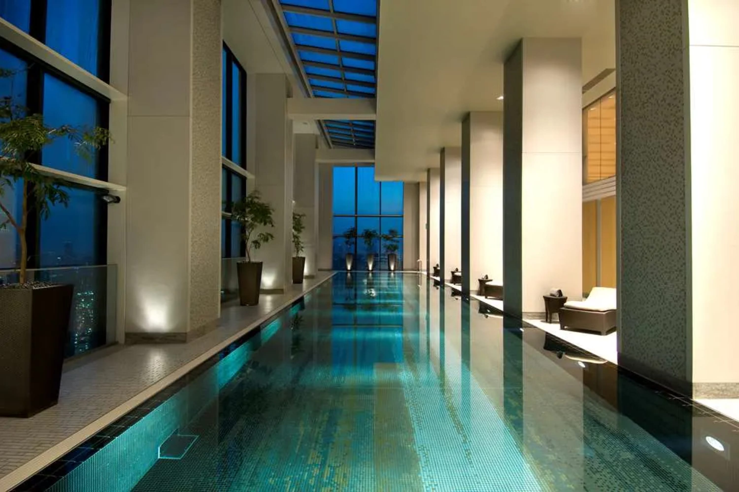 A lengthy indoor pool with an intricately tiled floor, surrounded by wide columns and views of Tokyo through large windows