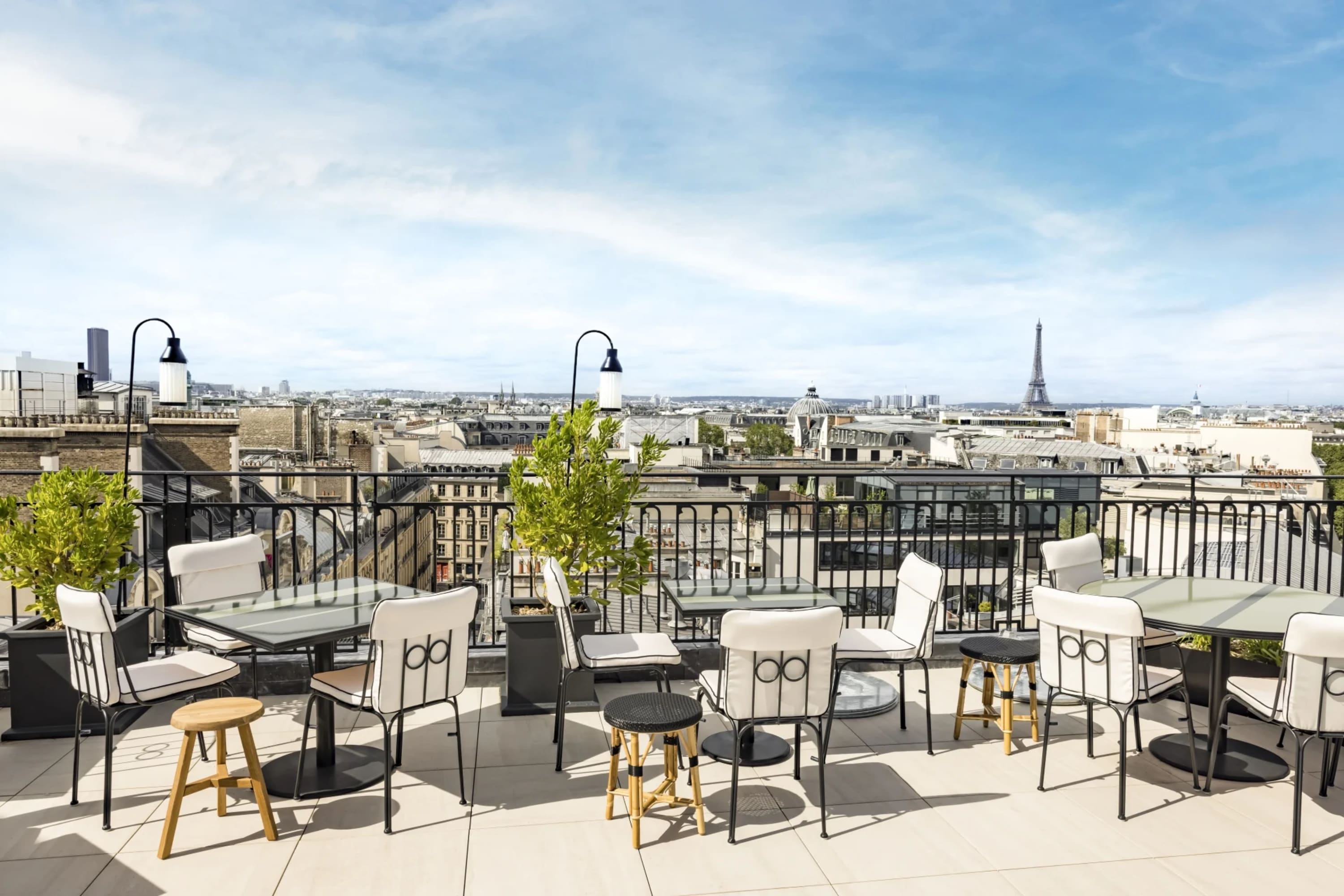 Trendy outdoor seating and tables buffeted by a wrought iron fence on a rooftop terrace overlooking the Paris skyline. The Eiffel Tower is visible in the far distance