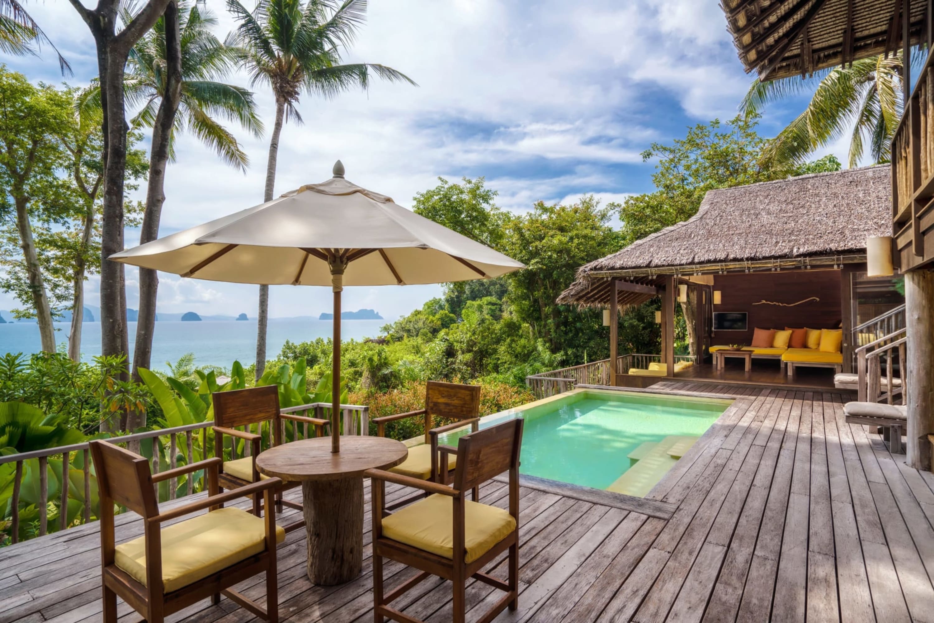 Rustic-esque island furniture on a wooden patio with a plunge pool. At the far side: outdoor furniture under a thatch roof